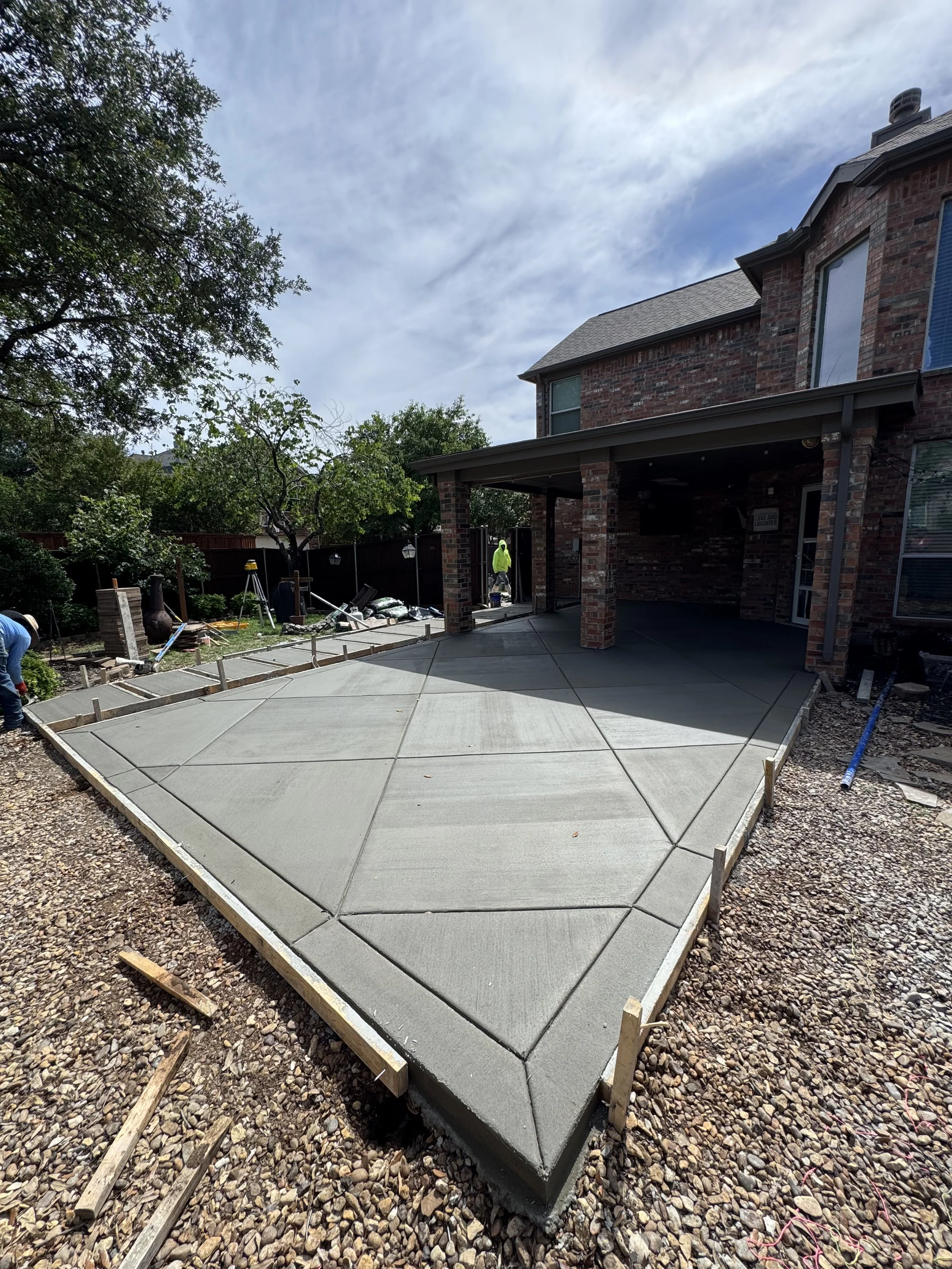 Newly poured concrete patio with formwork around edges, in a backyard with trees and a brick house, construction workers in safety gear working on the site.