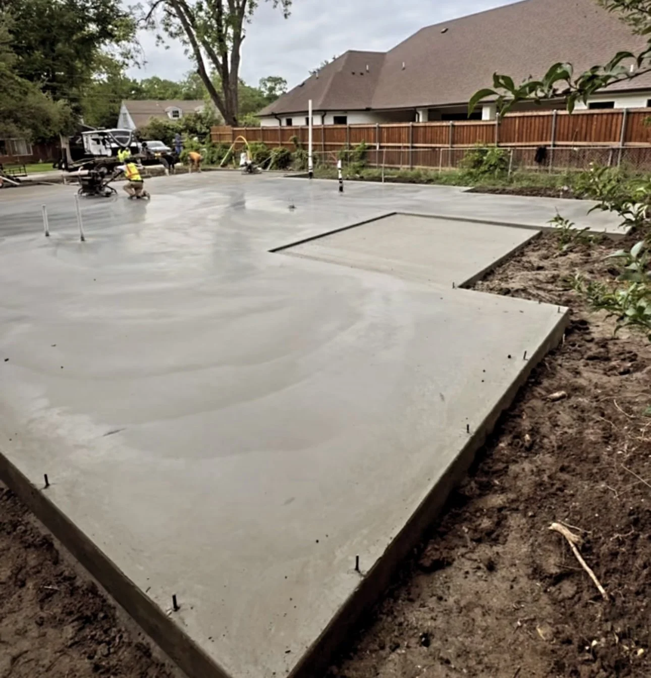 Newly poured concrete foundation slab in backyard with construction workers working in the background, wooden fence, trees, and houses.