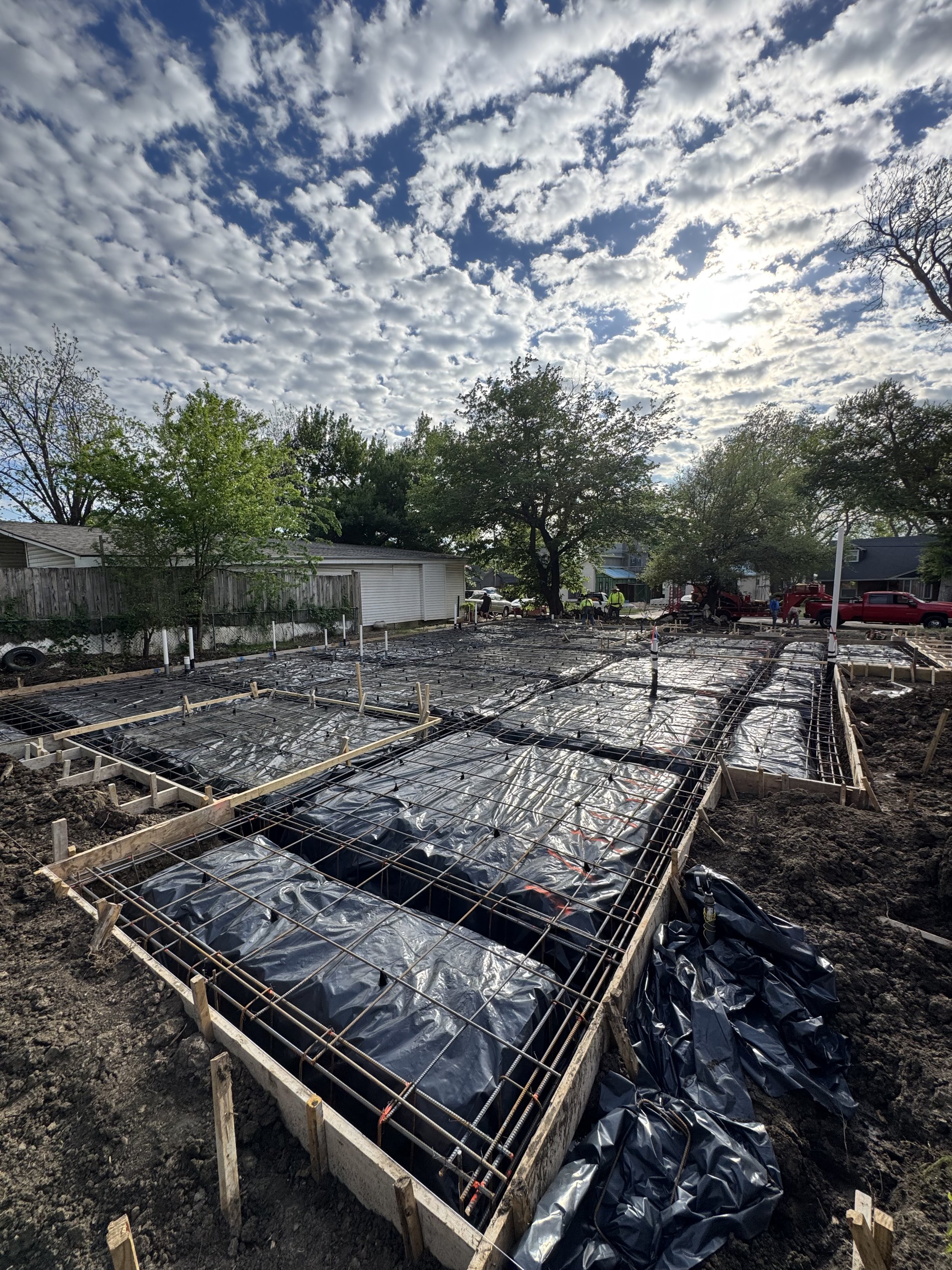 Construction site with freshly poured concrete foundation and steel rebar framing, surrounded by muddy ground and black plastic sheeting, with trees and residential houses in the background under a partly cloudy sky.