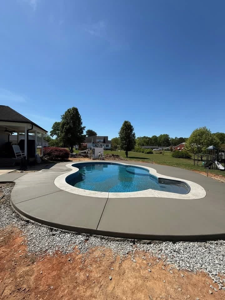 Backyard with a kidney-shaped swimming pool, concrete deck, and surrounding grass and trees under a clear blue sky.
