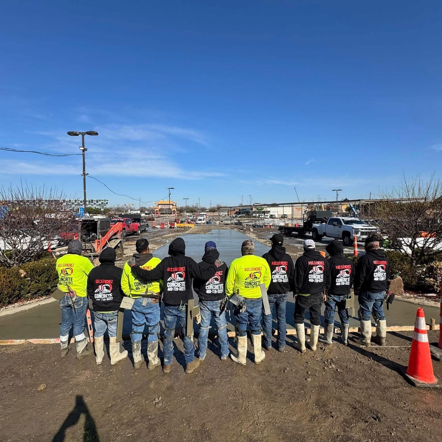 Group of construction workers wearing safety boots and bright jackets, observing a sidewalk construction site in a commercial parking lot on a sunny day.