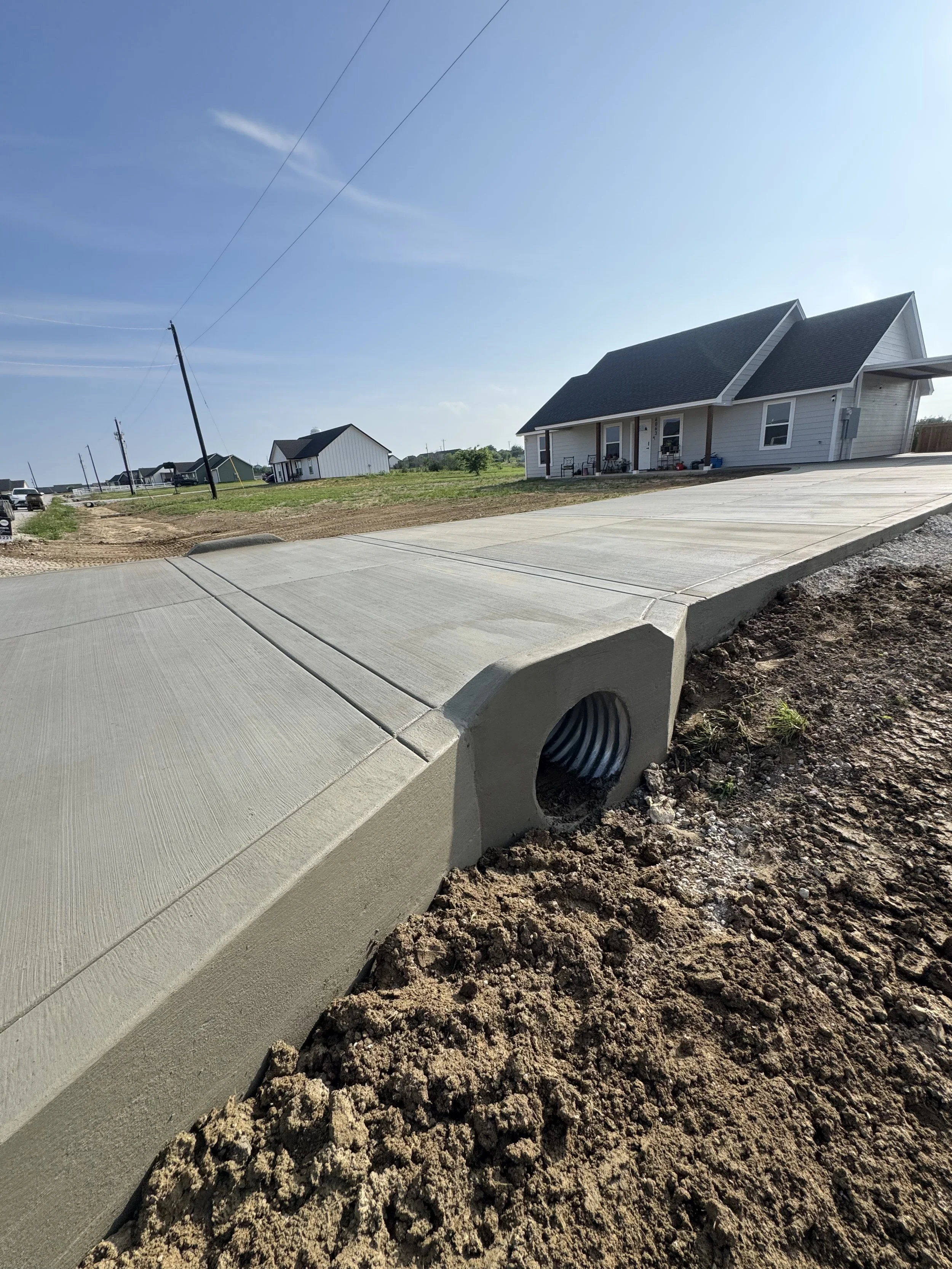 A newly poured concrete sidewalk in front of a white house with a black roof. The house has a small covered front porch with chairs and other items. Power lines run across the blue sky, and the surrounding area is mostly dirt and grass with other houses visible in the distance.