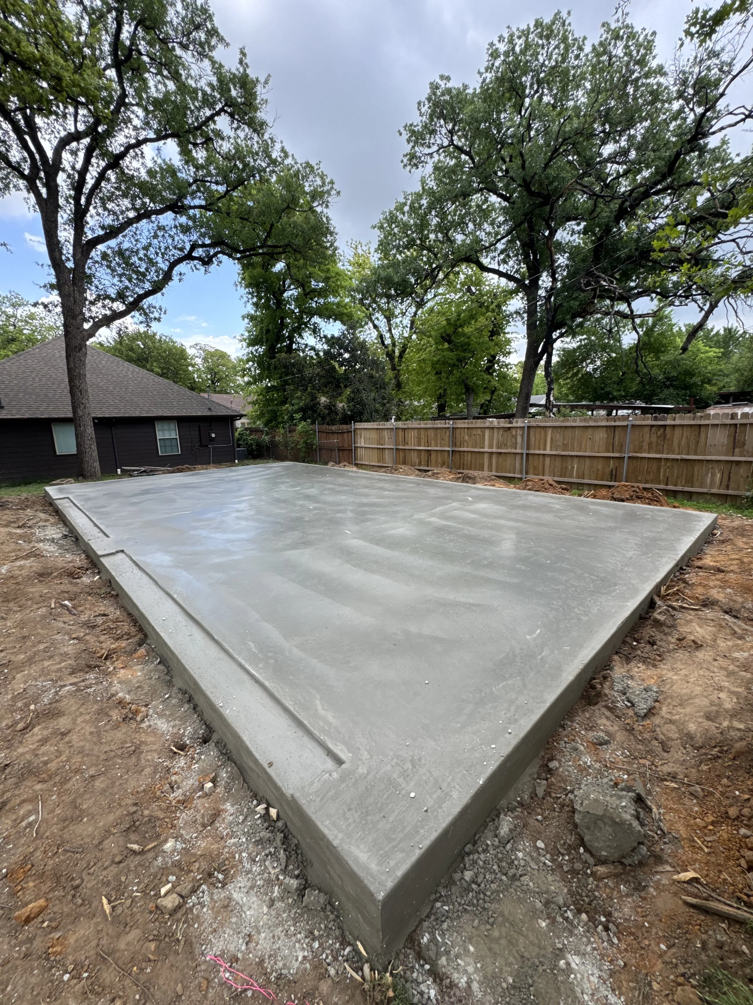 Freshly poured concrete slab in a backyard with a wooden fence, trees, a house, and a partly cloudy sky in the background.
