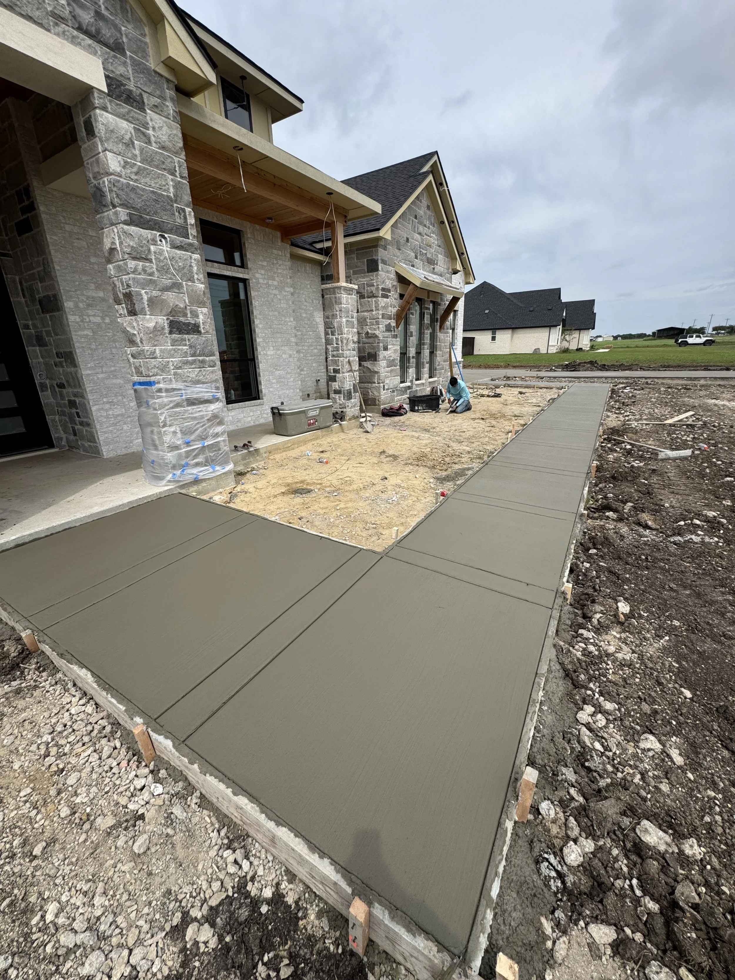 Concrete sidewalk being poured and finished outside a residential house under construction with brick exterior and a small porch overhang.