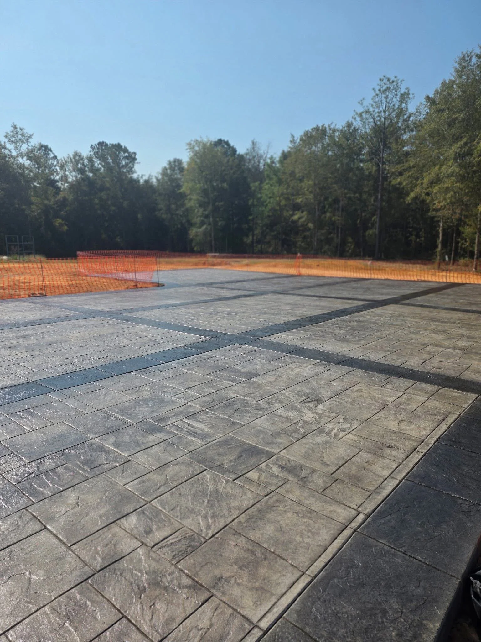 Newly paved outdoor area with decorative stone tiles, orange construction fencing, and a backdrop of trees and clear blue sky.