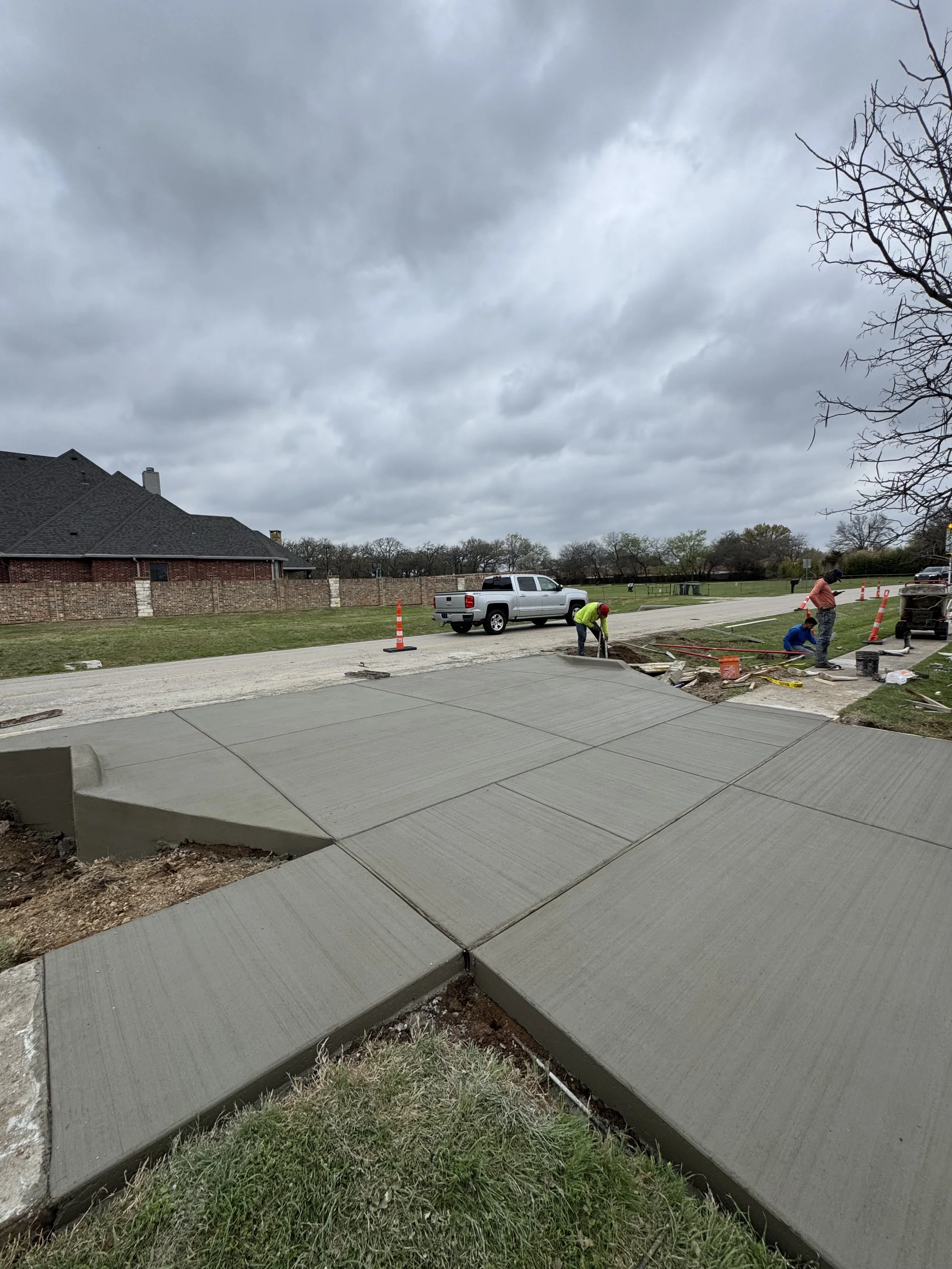 Workers installing a new concrete sidewalk, with some construction tools and cones nearby, under a cloudy sky in a residential neighborhood.