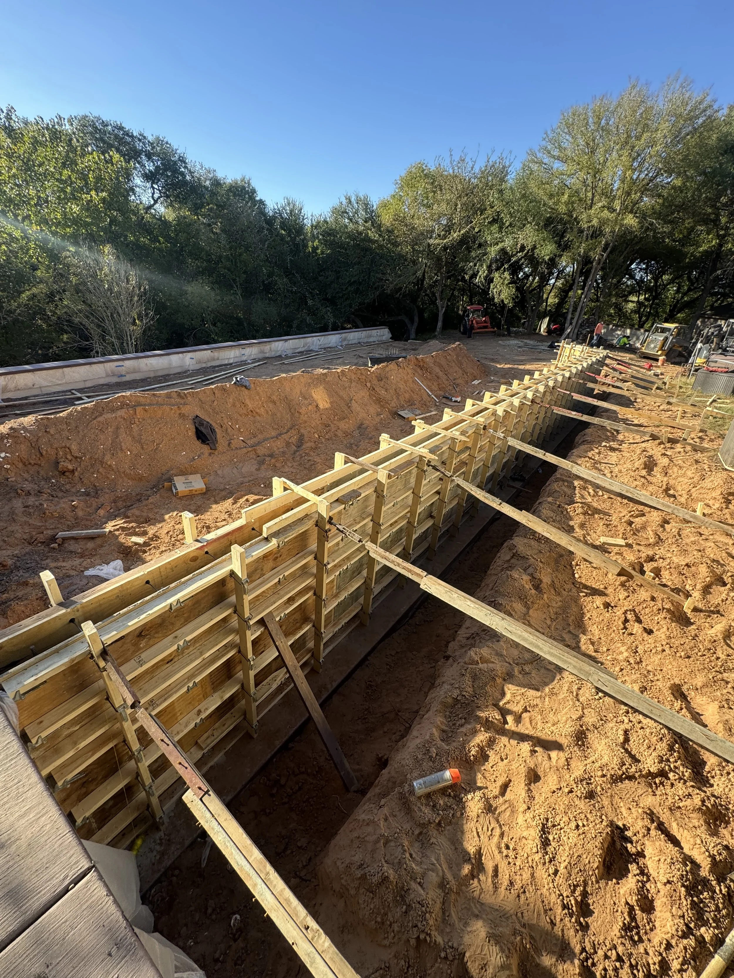 Construction site with wooden forms and trenches for building a new structure, surrounded by dirt and trees.