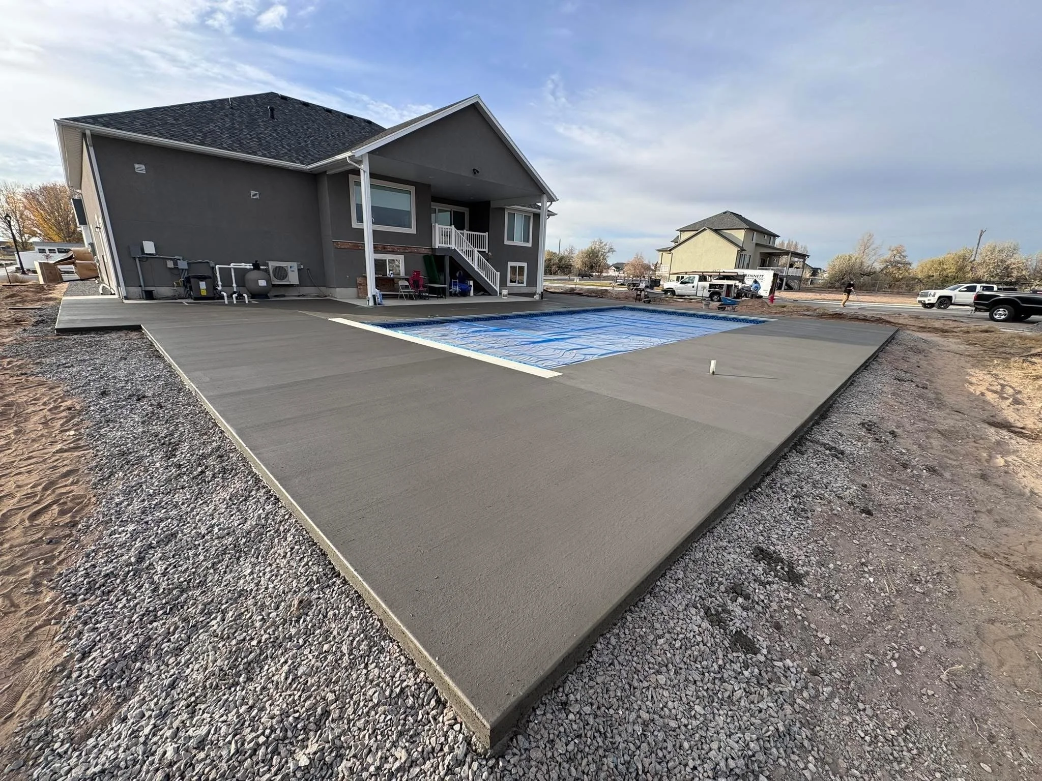 A newly poured concrete backyard with a covered patio and stairs leading to the second floor of a house, in a suburban neighborhood during daytime.