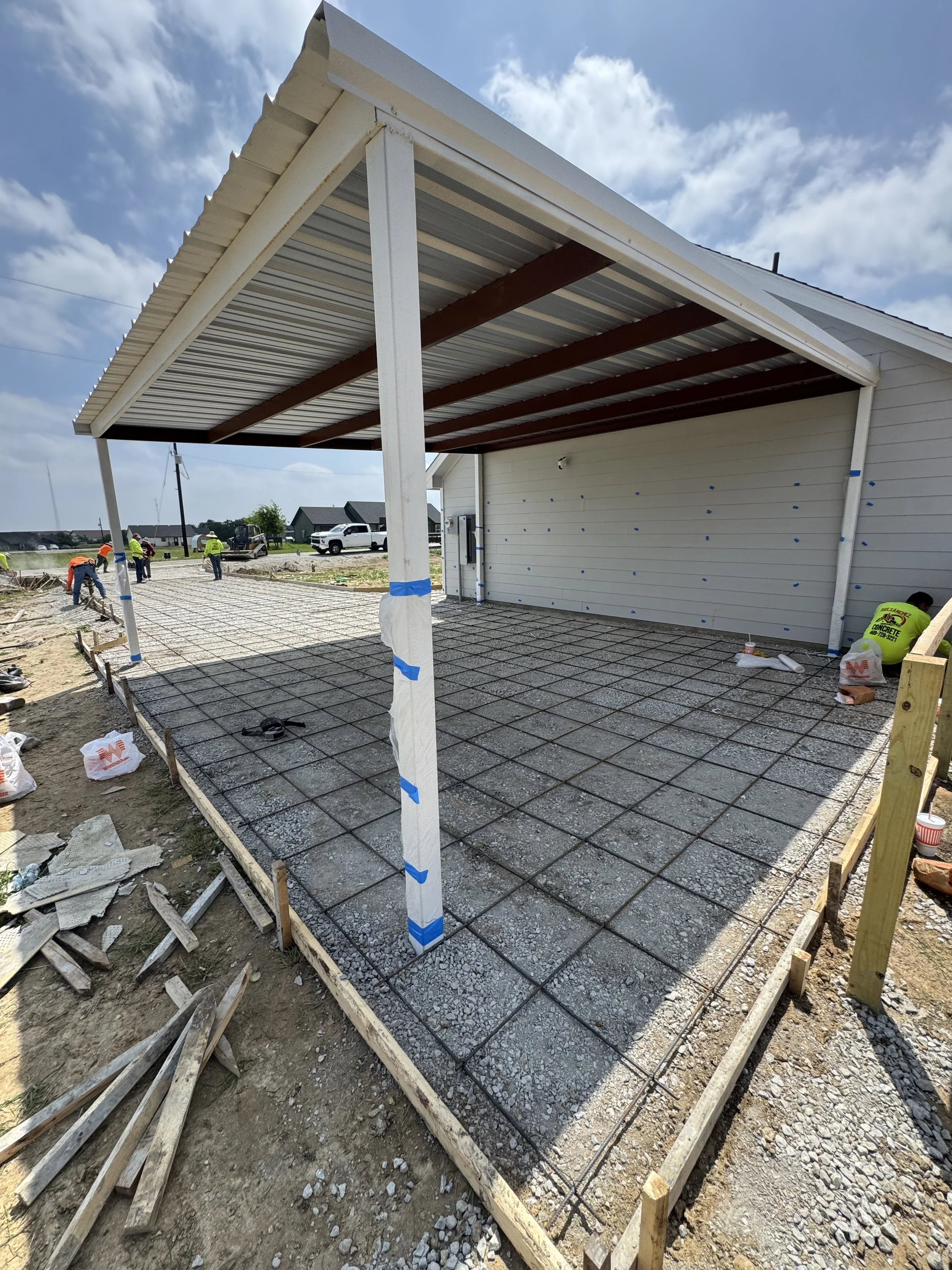 Construction site with workers installing concrete slab on a foundation with rebar grid, under a carport structure, in a suburban area with houses and a blue sky.