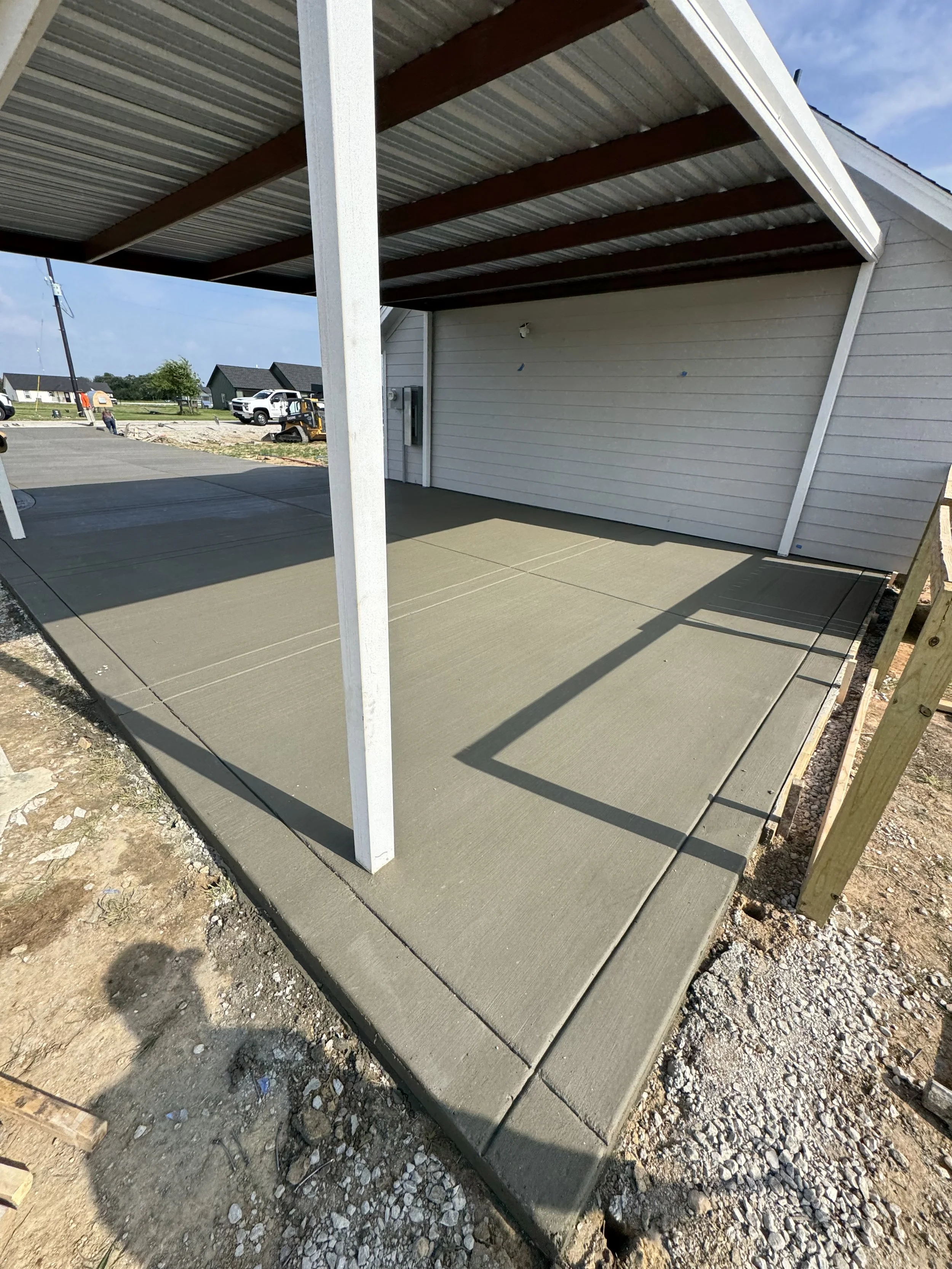 Newly poured concrete sidewalk under construction on a sunny day, with construction equipment and workers visible in the background.