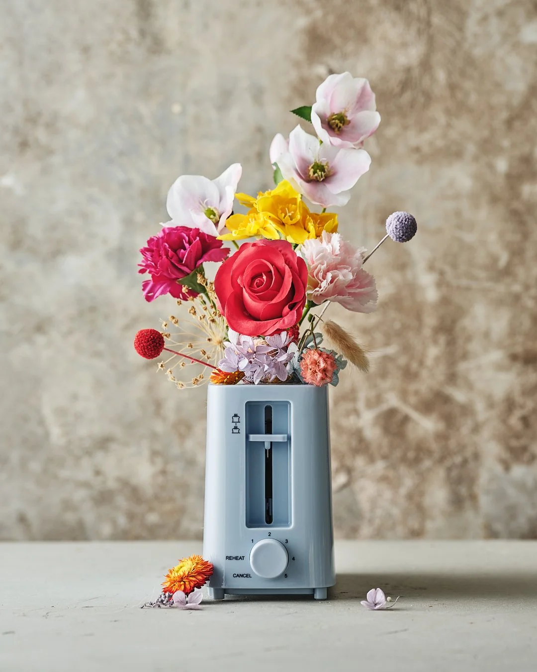 A white toaster with colorful flowers arranged in it, placed on a light surface against a textured beige background.