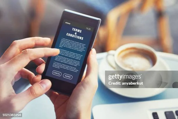 Person holding a smartphone with a terms and conditions agreement on the screen, sitting at a table with a coffee cup.