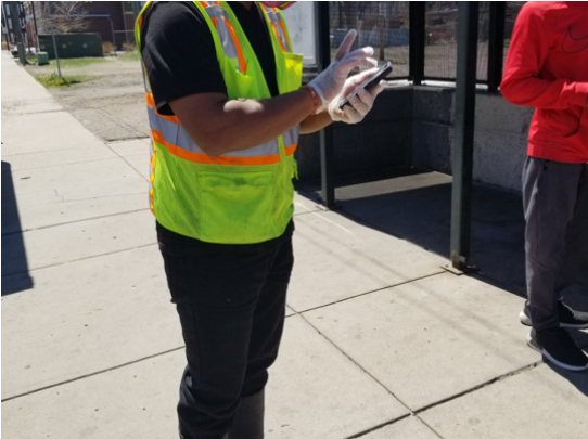 Person wearing a yellow reflector safety vest and gloves, standing outdoors on a sidewalk, looking at a smartphone.