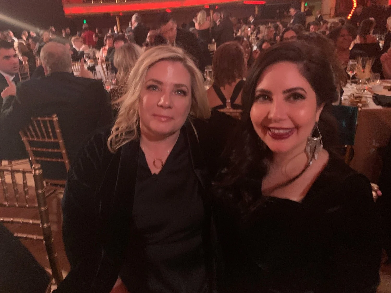 Two women sitting at a banquet table at a formal event, smiling at the camera. The background shows many other people seated at tables in a dimly lit venue.
