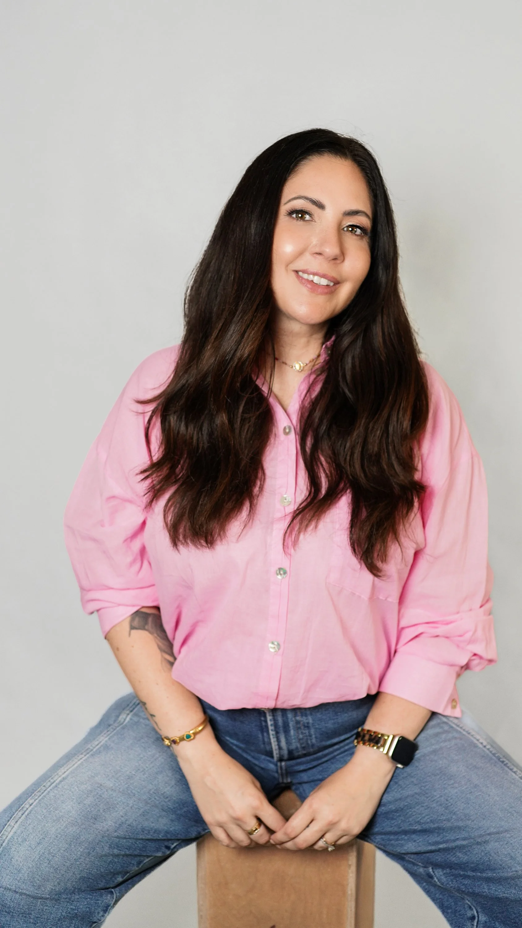 A woman with long dark hair, wearing a pink button-up shirt and blue jeans, sitting on a wooden box against a plain white background, smiling at the camera.