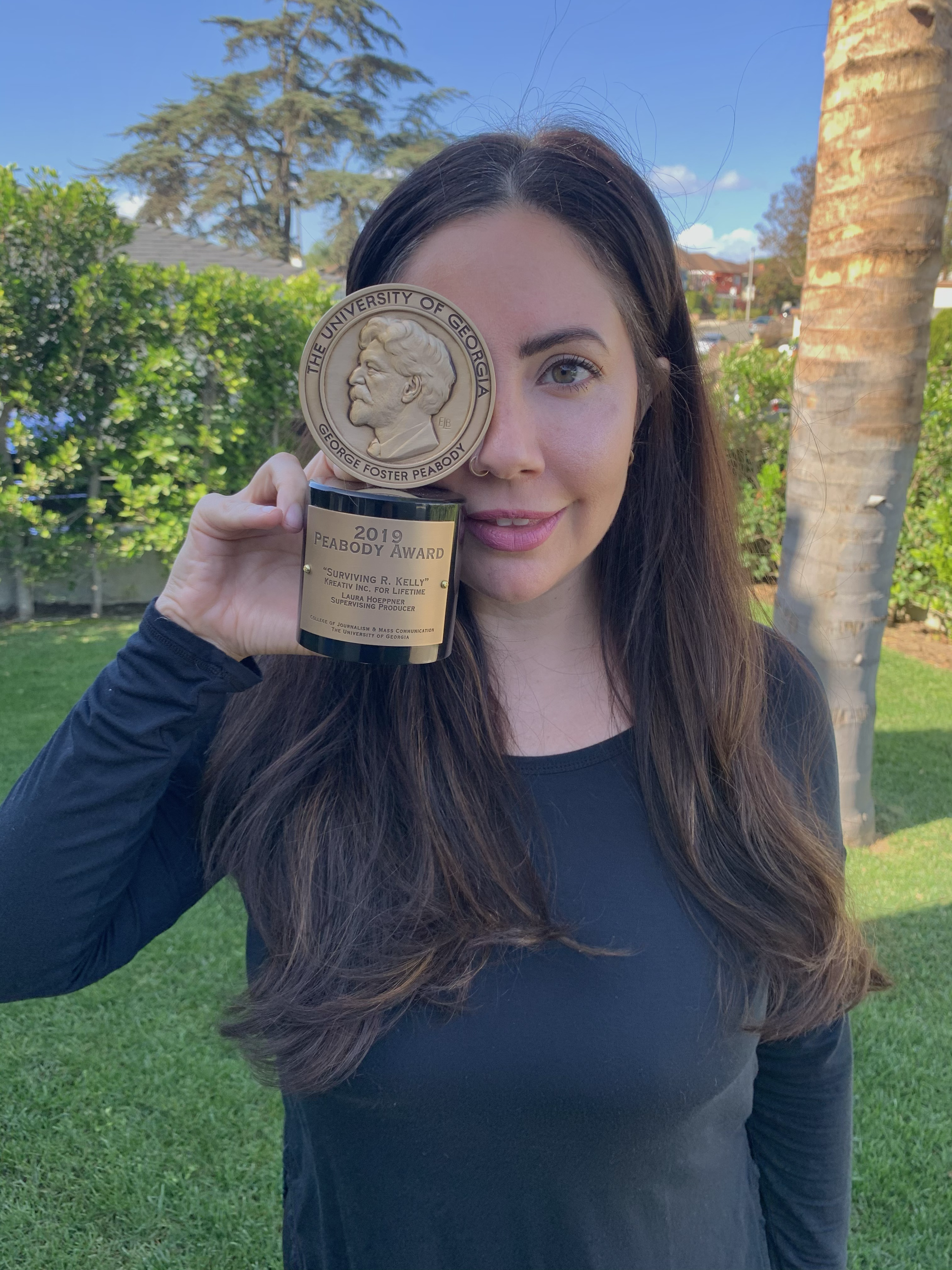 A woman with long brown hair and fair skin holding an award plaque outdoors. The award features a large medallion with a portrait of George Foster Peabody from the University of Georgia, and a black base with a gold plate stating 2019 Peabody Award f