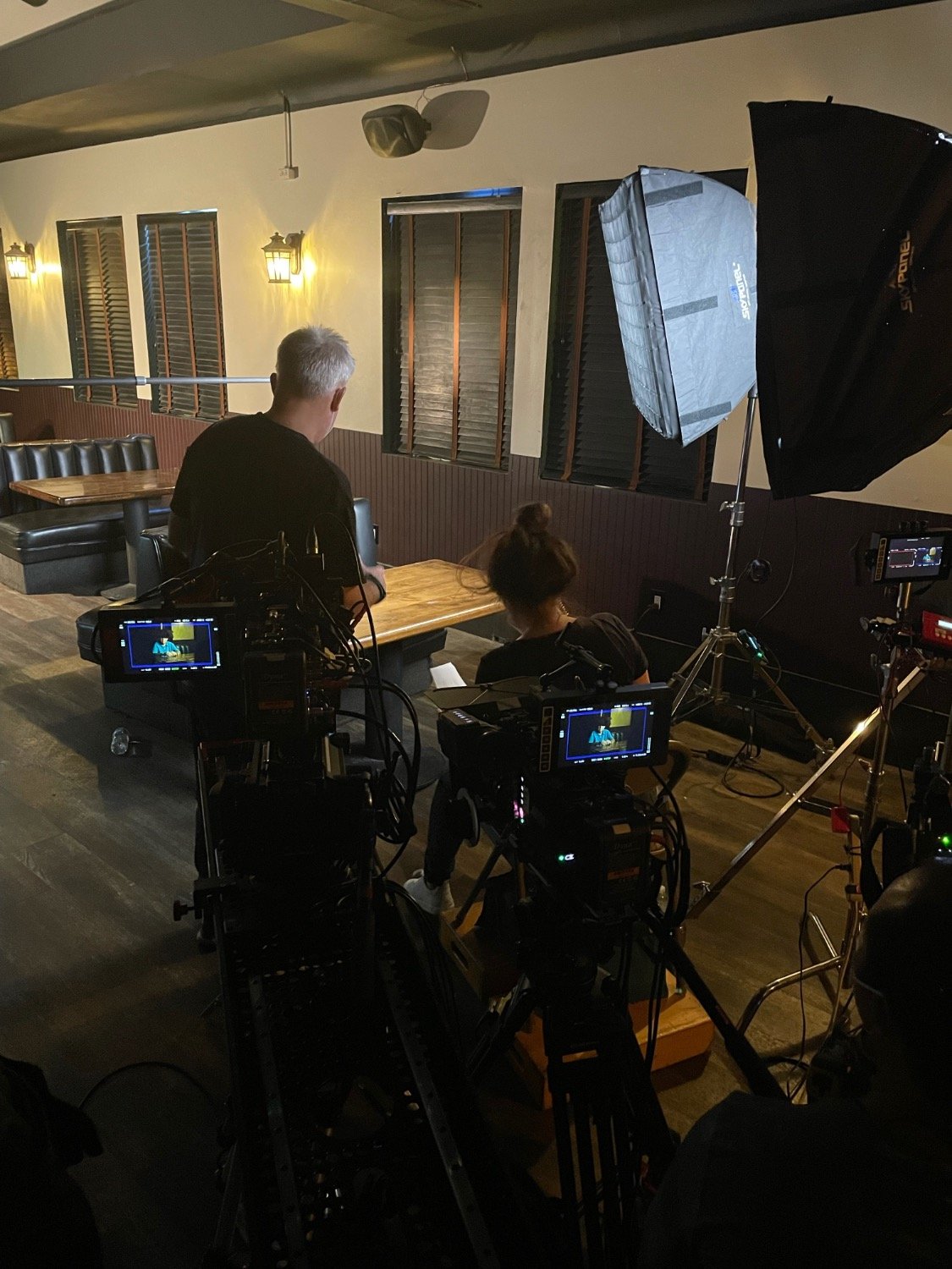 Inside a recording studio, two crew members operate cameras and lighting equipment while a woman sits at a wooden table in front of the camera setup.