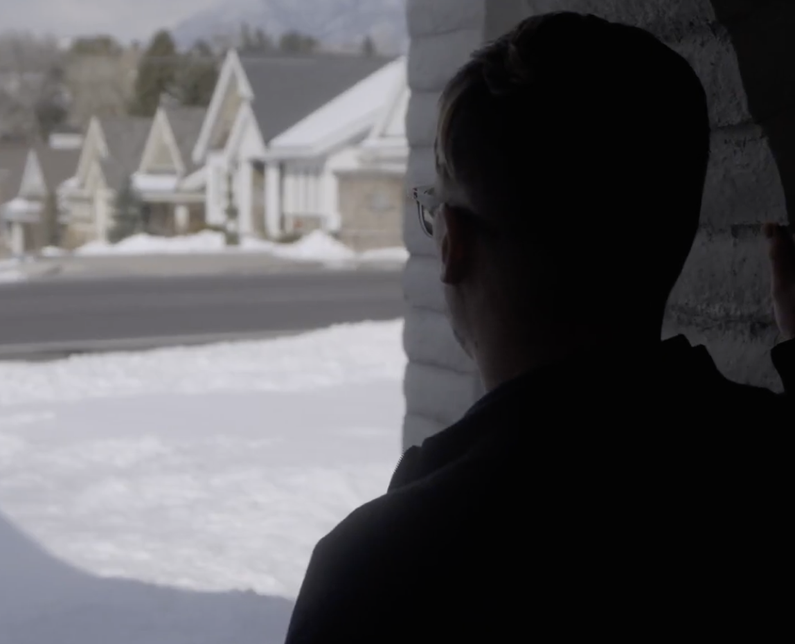 A person with glasses looking out from inside a house through a window at a snowy suburban neighborhood.