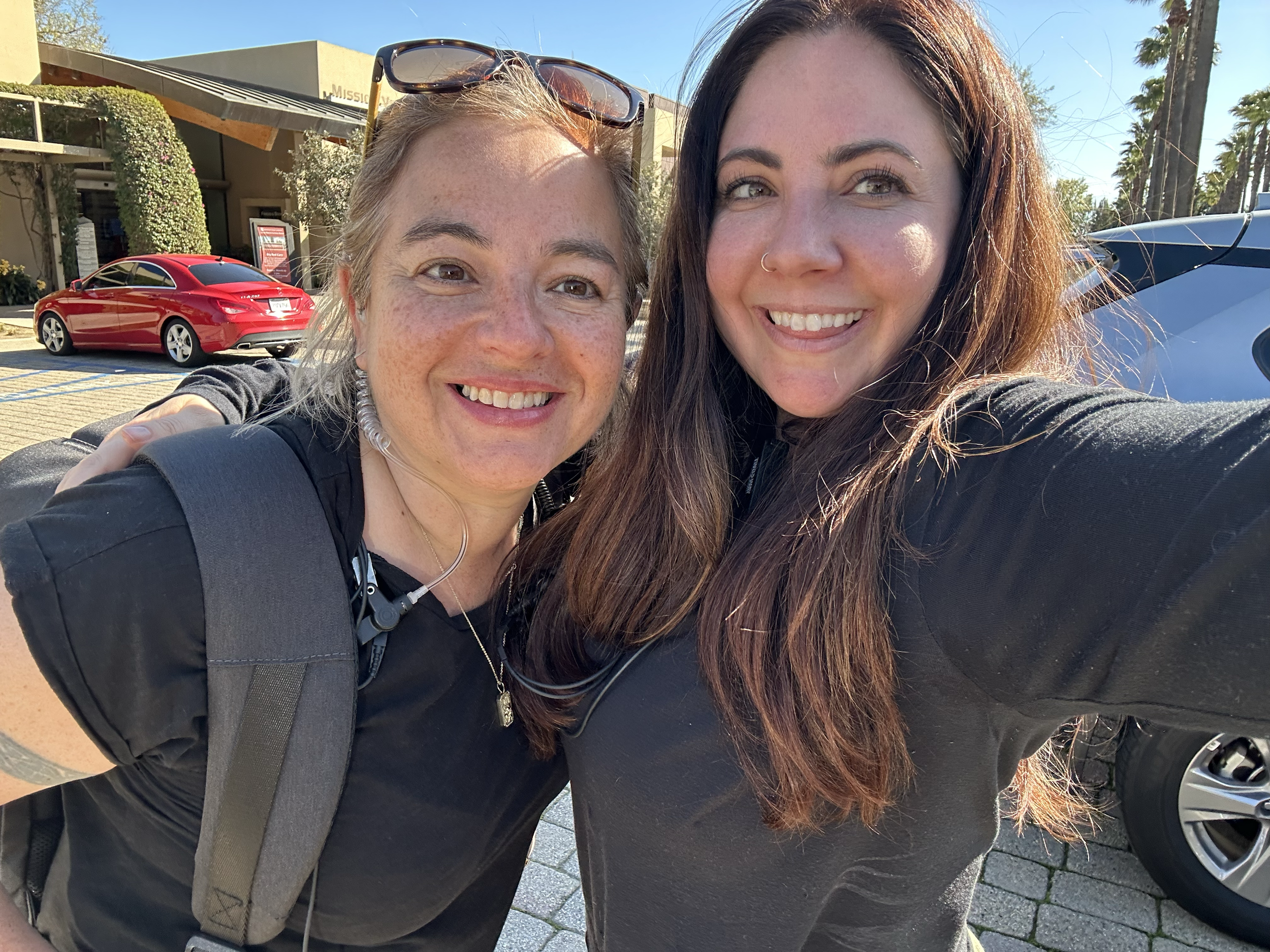 Two women smiling for a selfie outdoors on a sunny day. One has light skin, freckles, glasses on her head, and a necklace. The other has light skin, long dark hair, and a nose ring. In the background, there is a red car, palm trees, and a building.