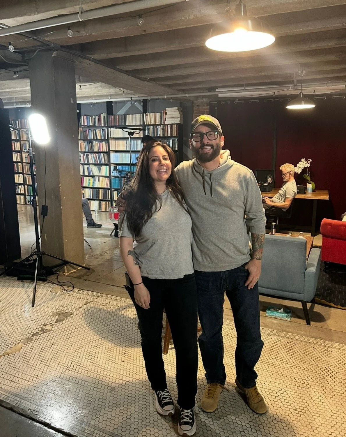 Two smiling people standing close together in a cozy, stylish indoor space with bookshelves, furniture, and a person working at a desk in the background.