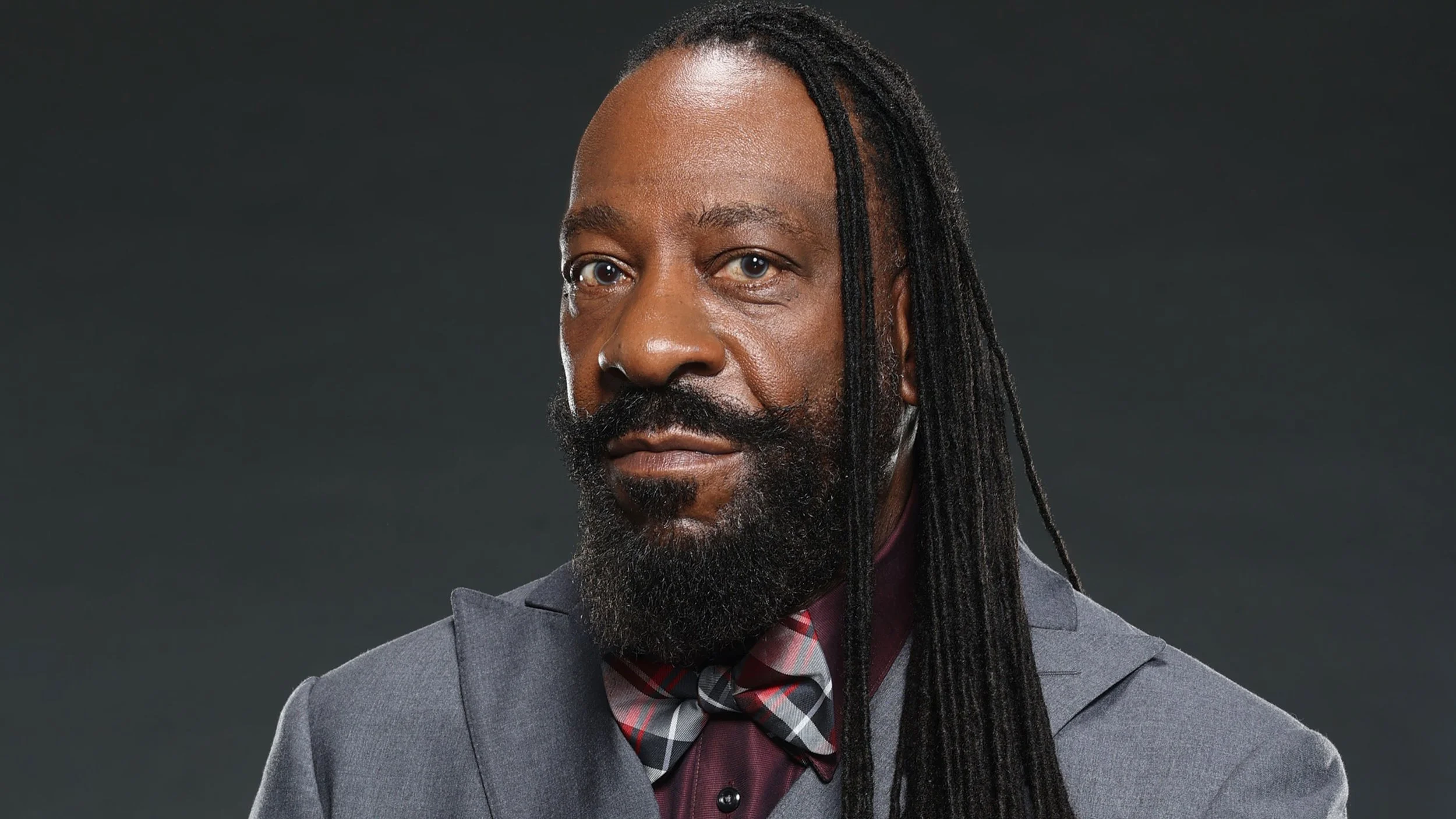 A man with a beard and long dreadlocks, dressed in a suit and a plaid bowtie, posing against a dark background.