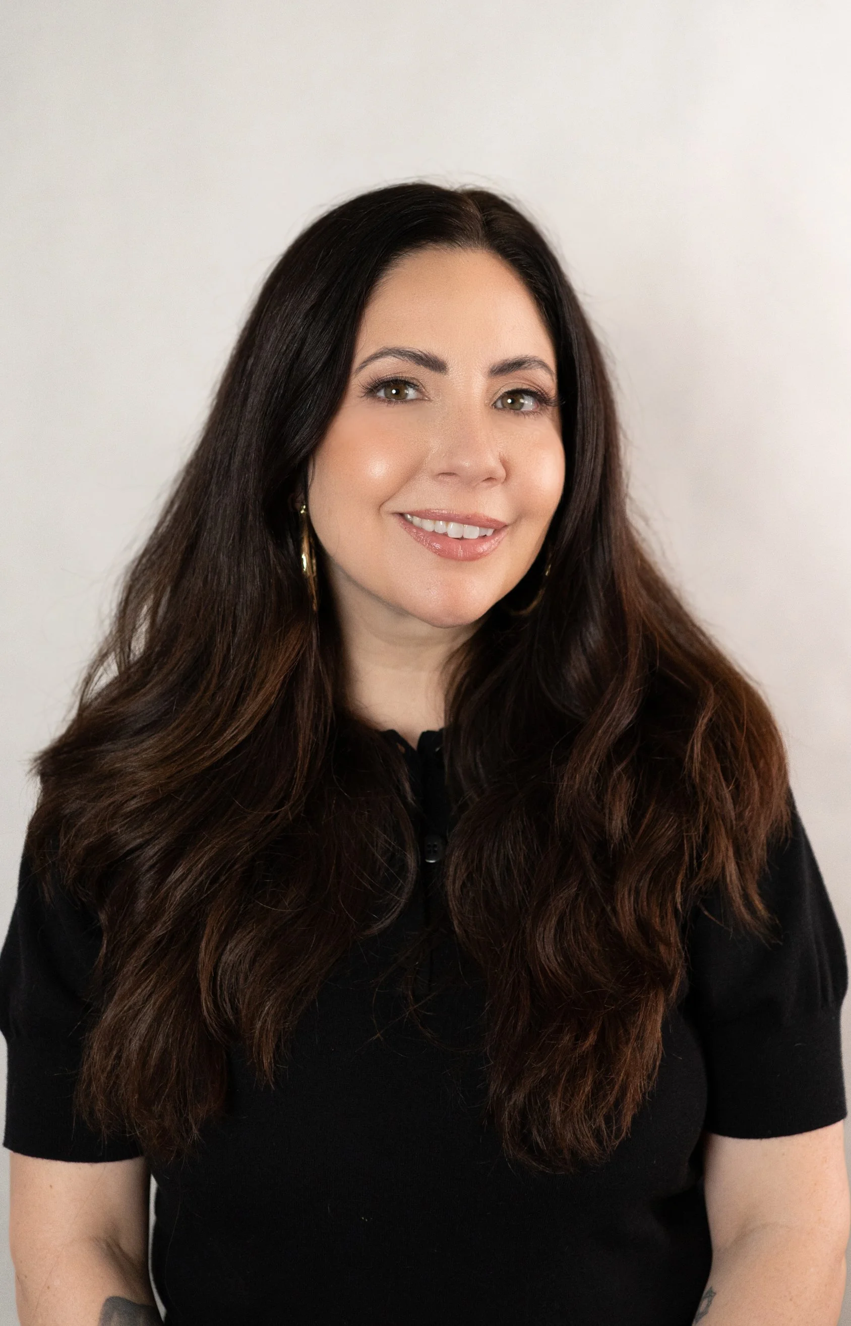 A woman with long, wavy brown hair and light skin, smiling, wearing gold hoop earrings and a black top, standing against a plain white background.