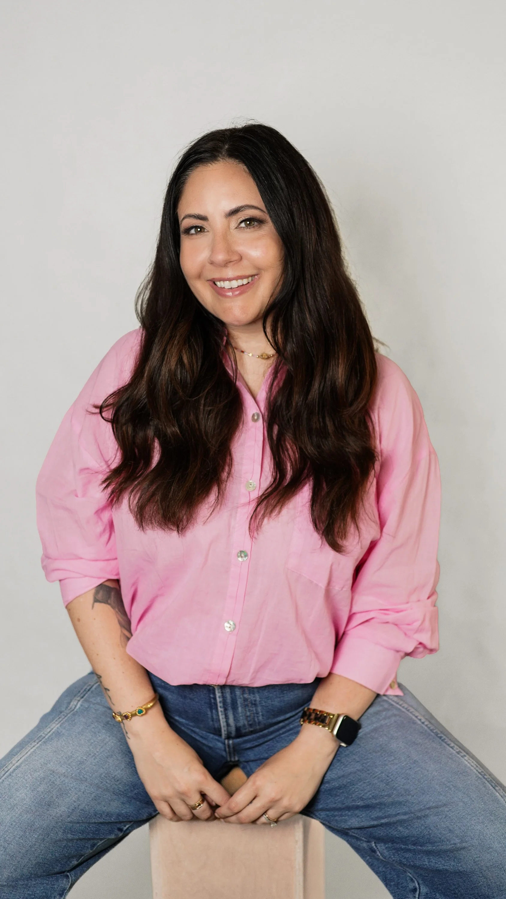 A woman with long dark hair sitting on a wooden block, wearing a pink button-up shirt and blue jeans, smiling at the camera against a plain light-colored background.
