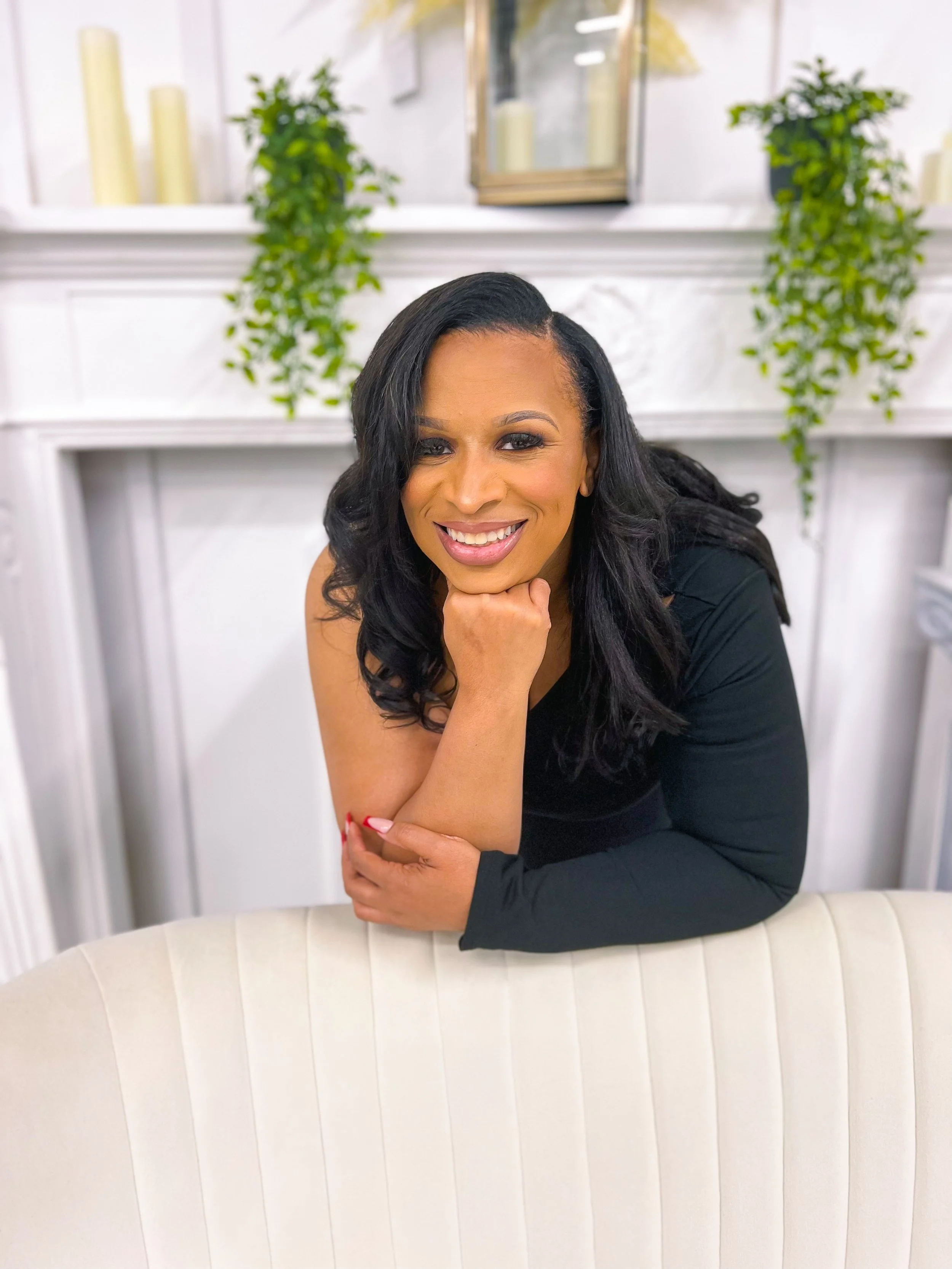 A woman with long dark hair smiling, resting her chin on her hand, sitting in front of a white fireplace with green plants on the mantel.
