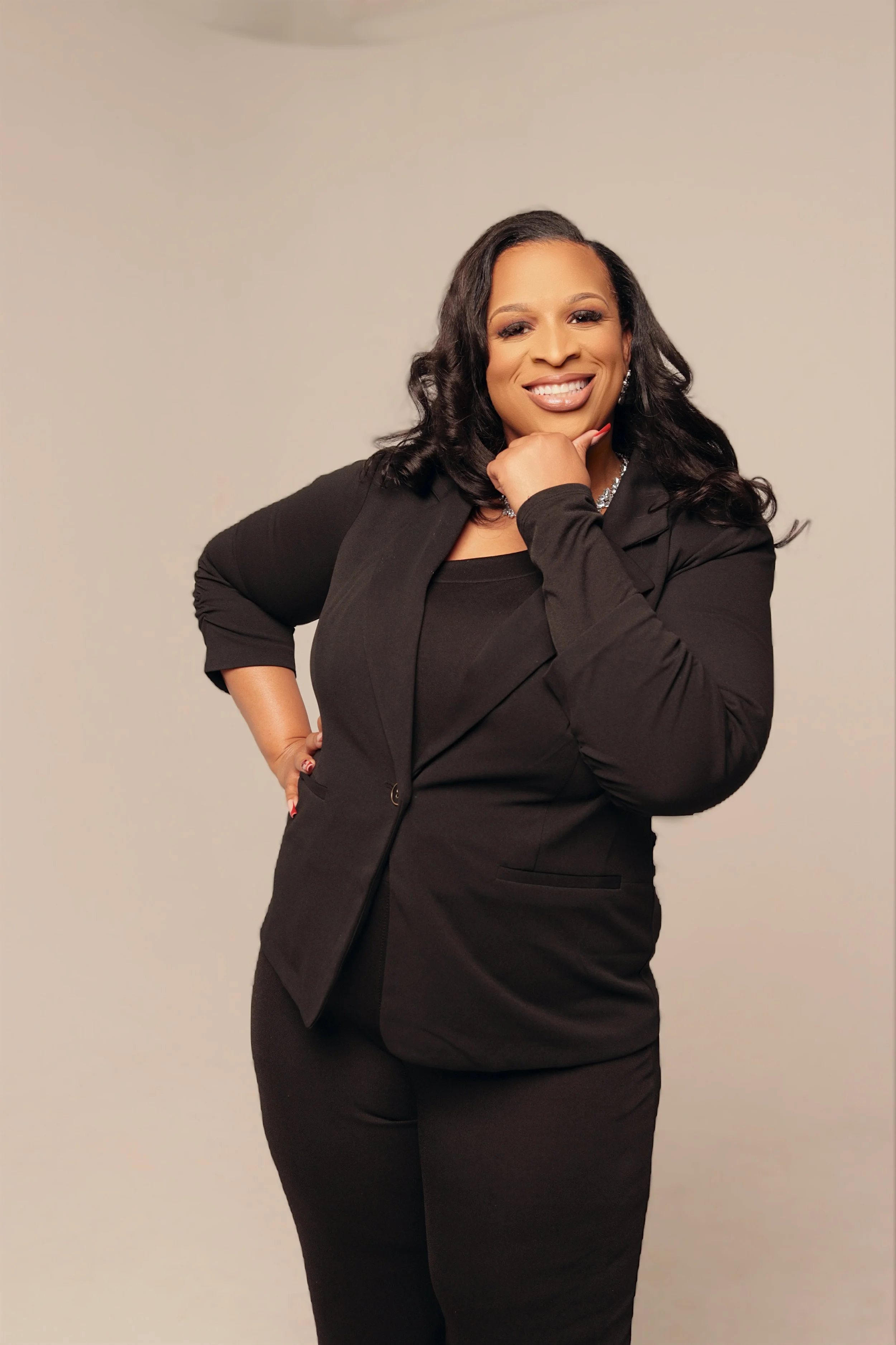 Woman with black hair, wearing a black blazer and black skirt, smiling with one hand resting on her chin against a plain light-colored background.