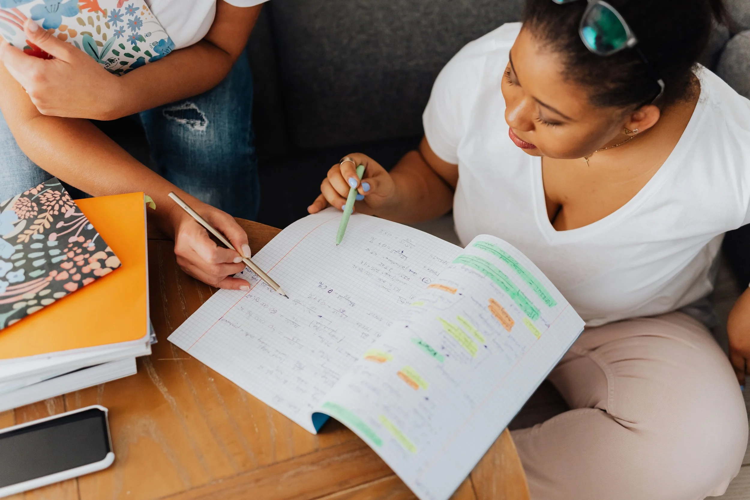 A woman with dark curly hair and sunglasses on her head is sitting at a wooden table, writing in a notebook with handwritten notes and highlighting. Next to her are stacked notebooks and a smartphone.