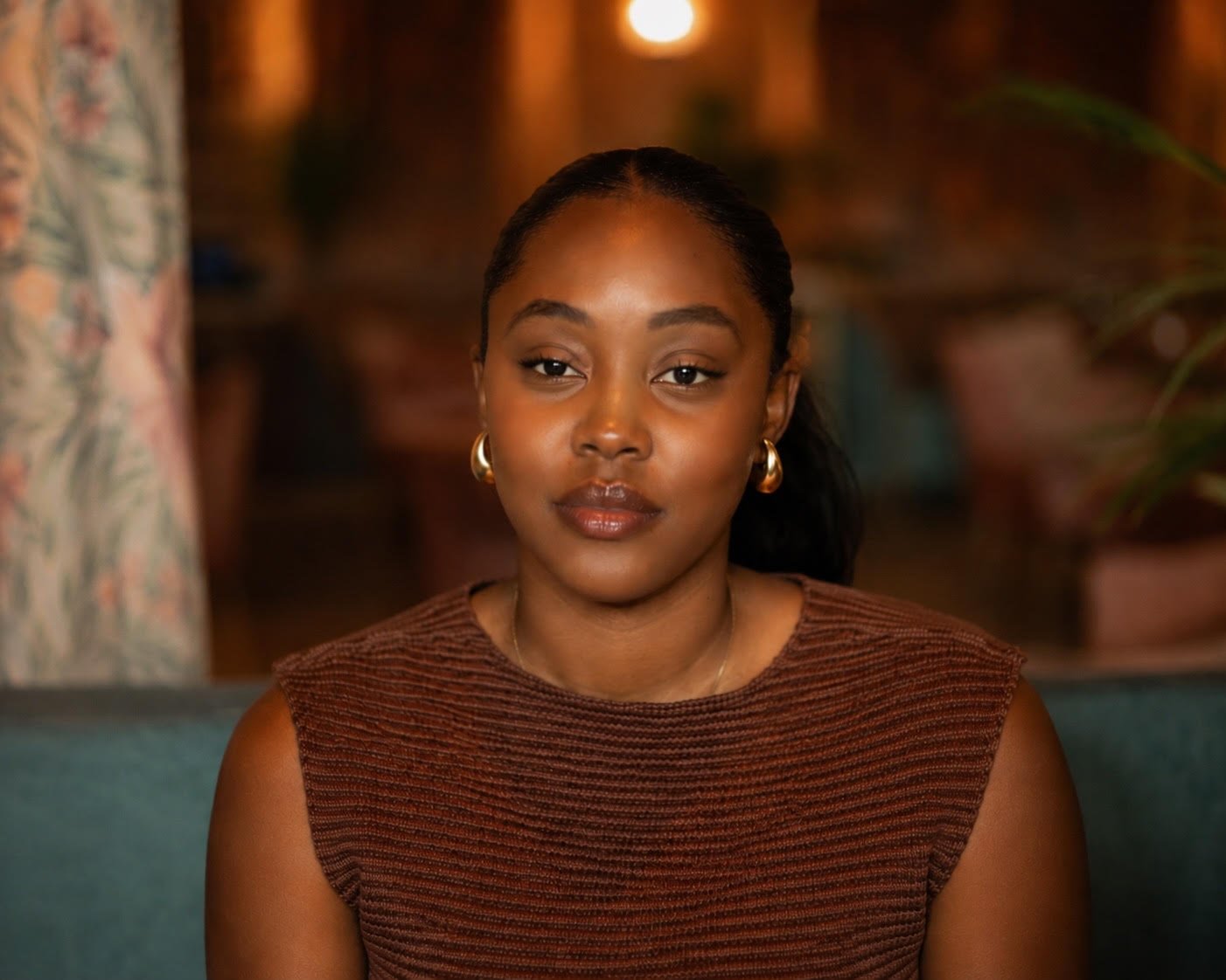 A woman with dark hair tied back, wearing gold hoop earrings and a sleeveless rust-colored top, sitting indoors against a dimly lit background with warm tones. Noah Peters