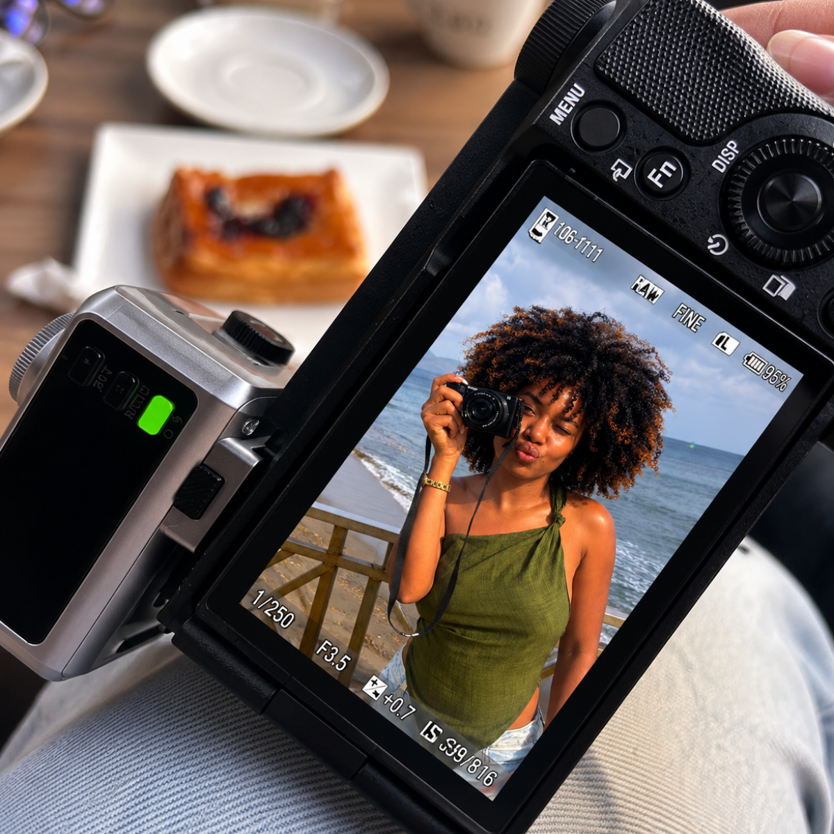 Une femme souriante avec des cheveux bouclés pose avec un appareil photo sur une plage, visible à travers l'écran d'un appareil photo numérique. En arrière-plan, des croissants et des tasses sont posés sur une table en bois.