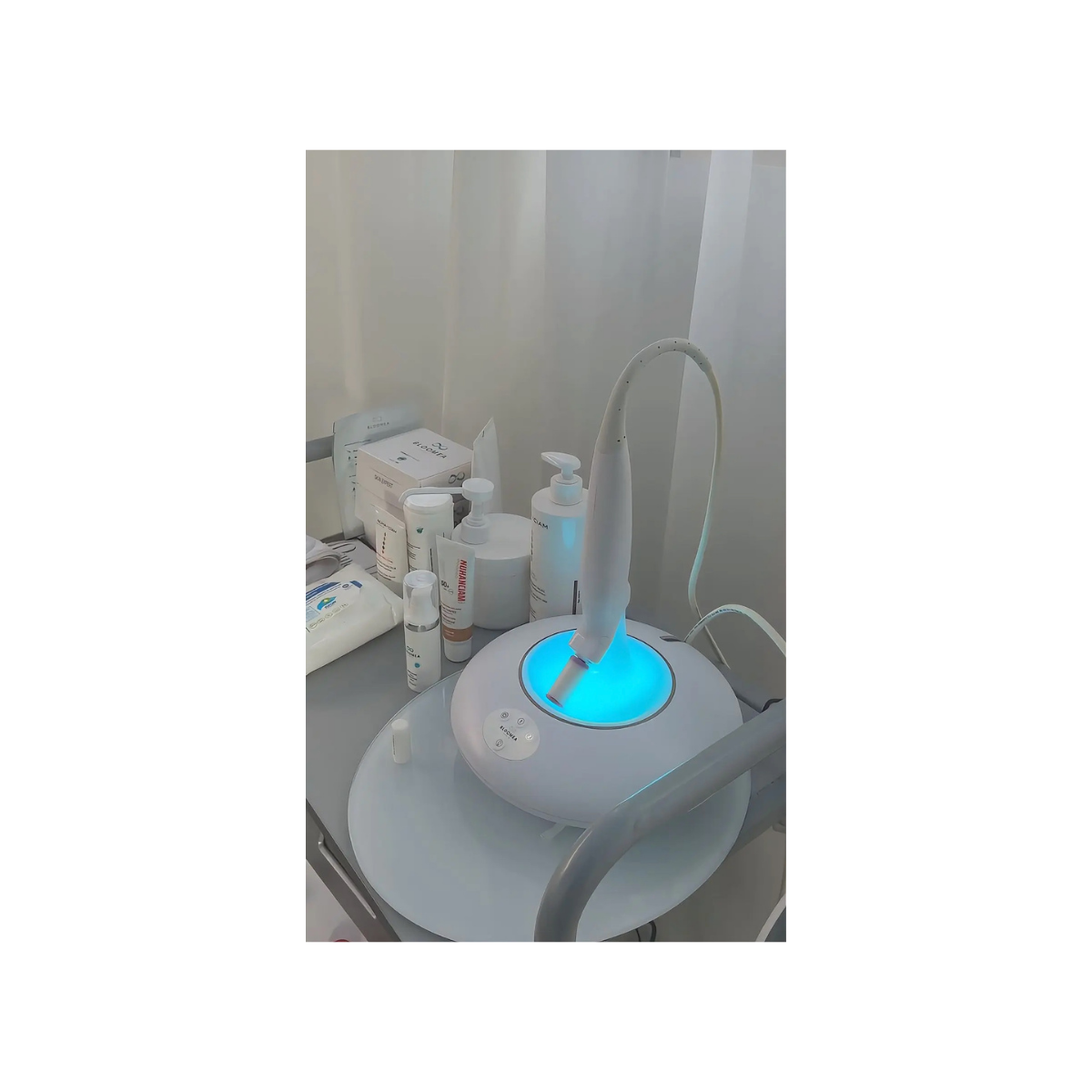 Un appareil de nettoyage facial avec une lumière bleue, placé sur un meuble avec des produits de soin, dans un environnement clinique ou esthétique.