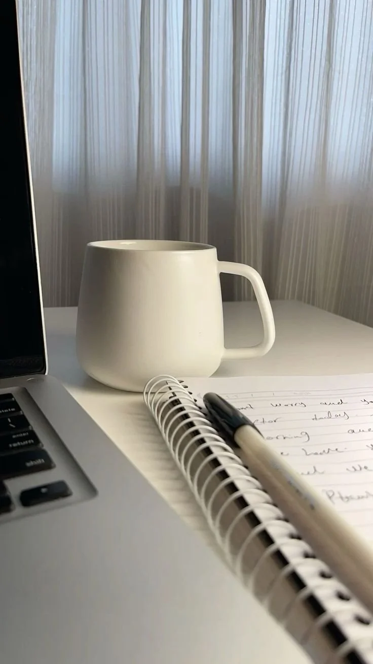 Tasse blanche, ordinateur portable, carnet et stylo sur un bureau, avec des rideaux en arrière-plan.