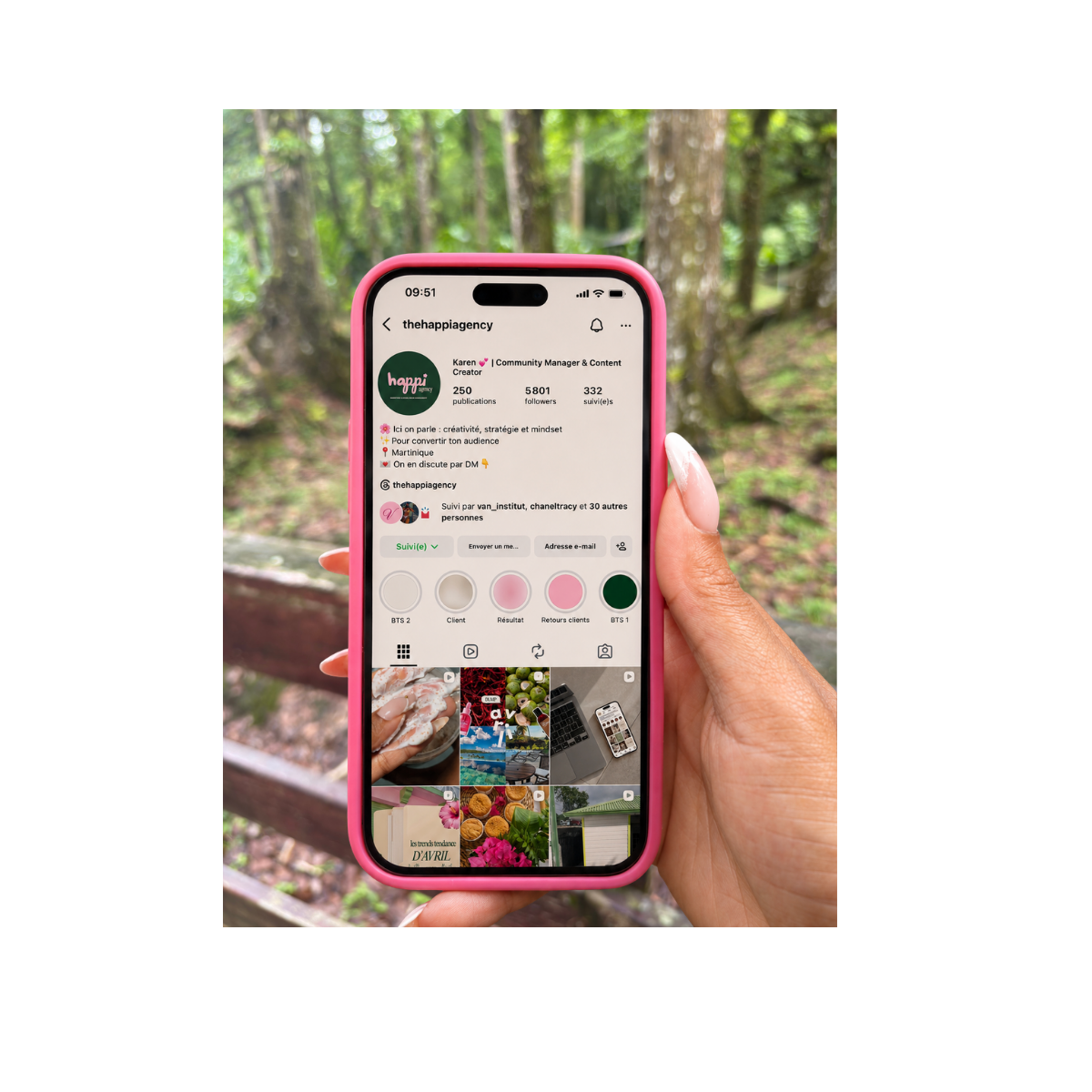 Une personne tient un téléphone avec un étui rose, affichant un profil Instagram d'une agence de communication. Le fond est une forêt verdoyante.