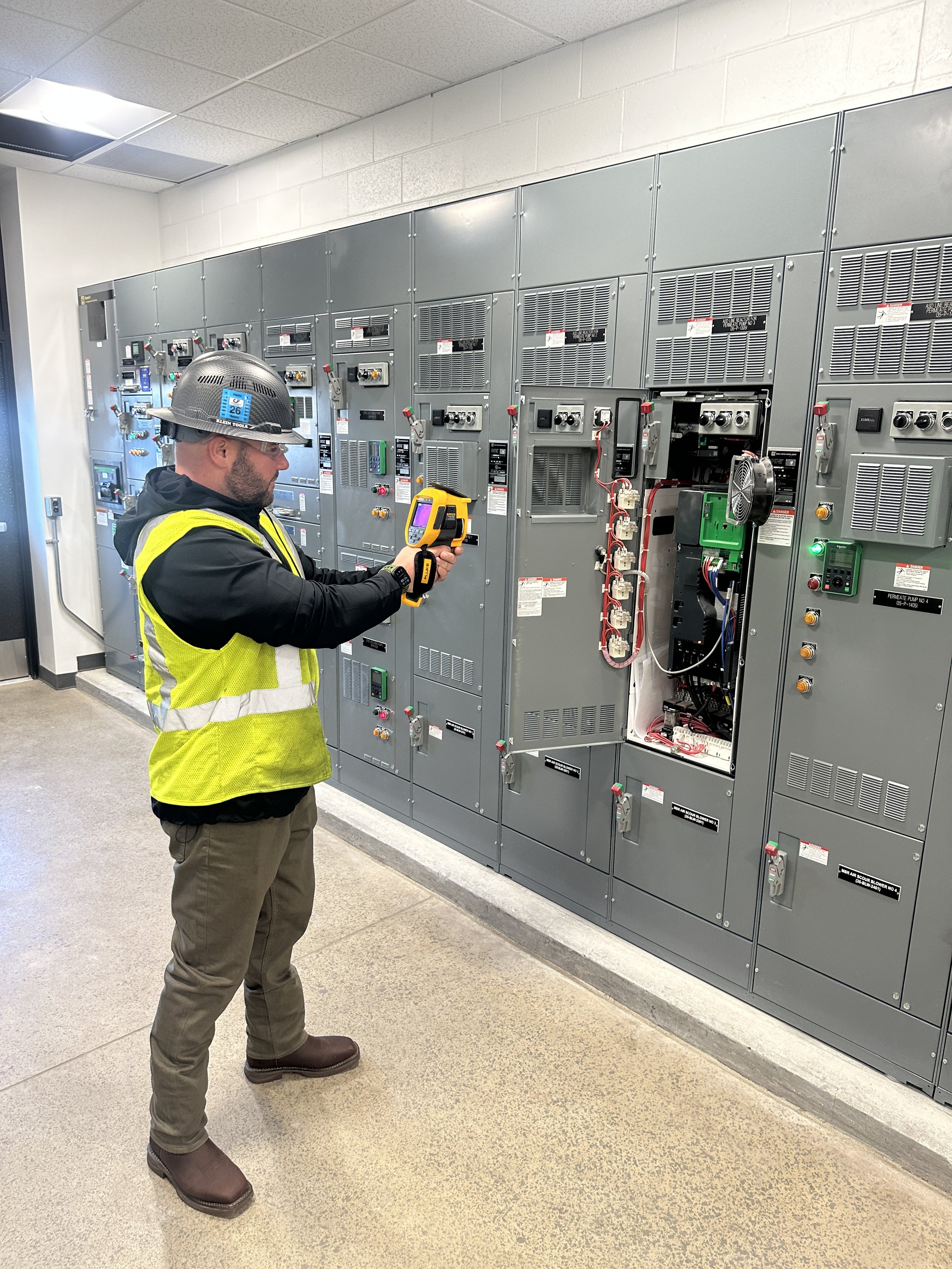 A man wearing a safety helmet, glasses, and a yellow vest is inspecting electrical equipment in a room with industrial electrical panels.
