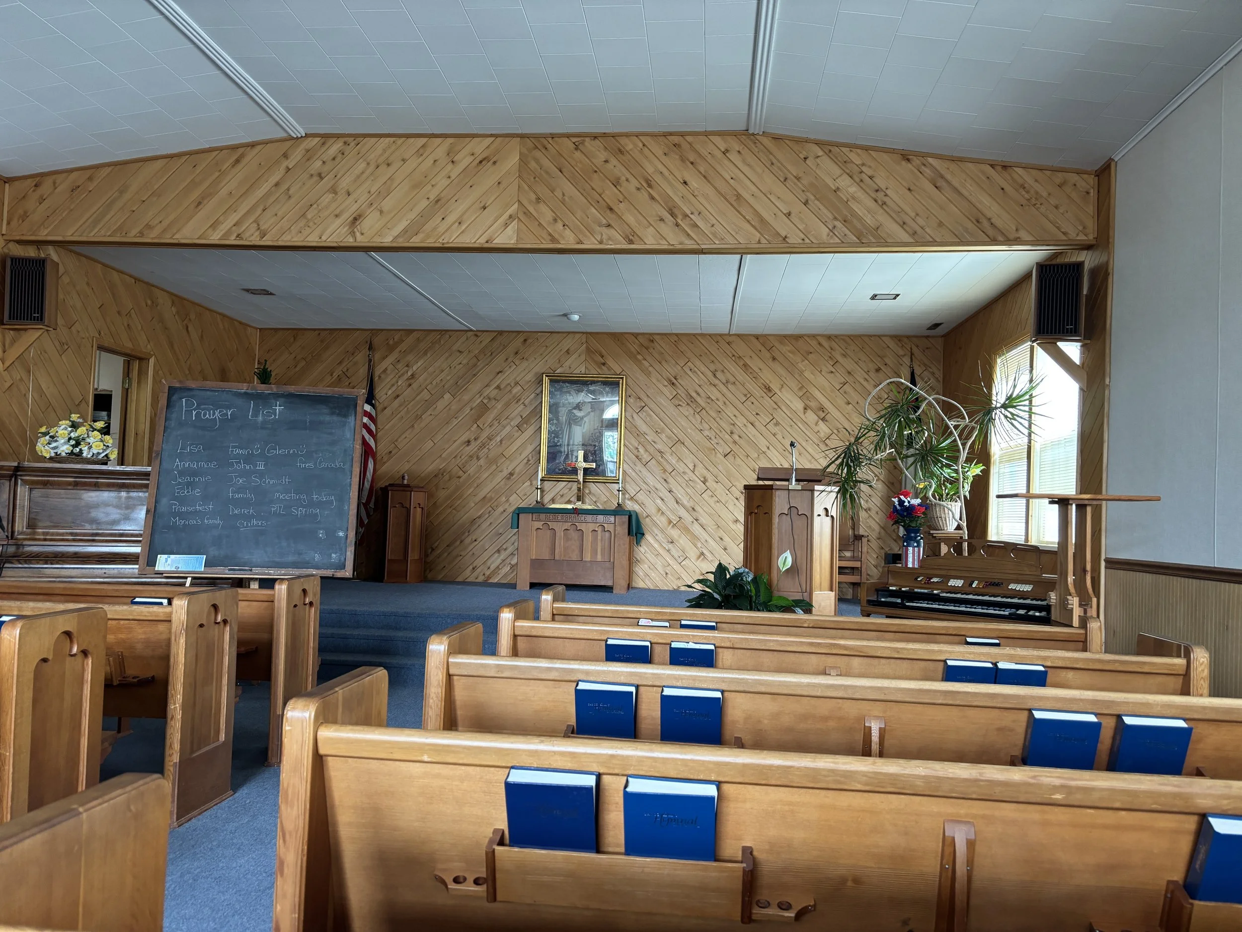 Inside of a church with wooden pews, a chalkboard with a prayer list, and a small raised stage at the front with religious items and decorations.