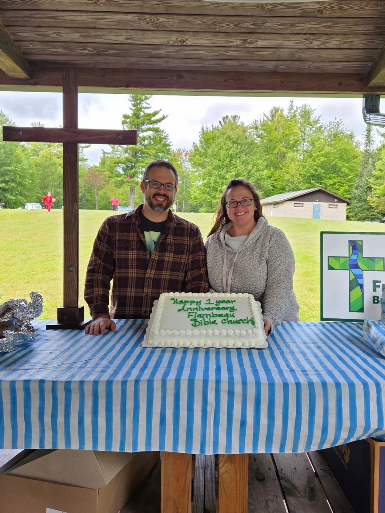 Two people, a man and a woman, smiling behind a birthday cake at an outdoor celebration at Flambreau Bible Church. The cake has green writing that reads 'Happy 1 Year Anniversary, Flambreau Bible Church.' There is a wooden cross and a church logo on display.
