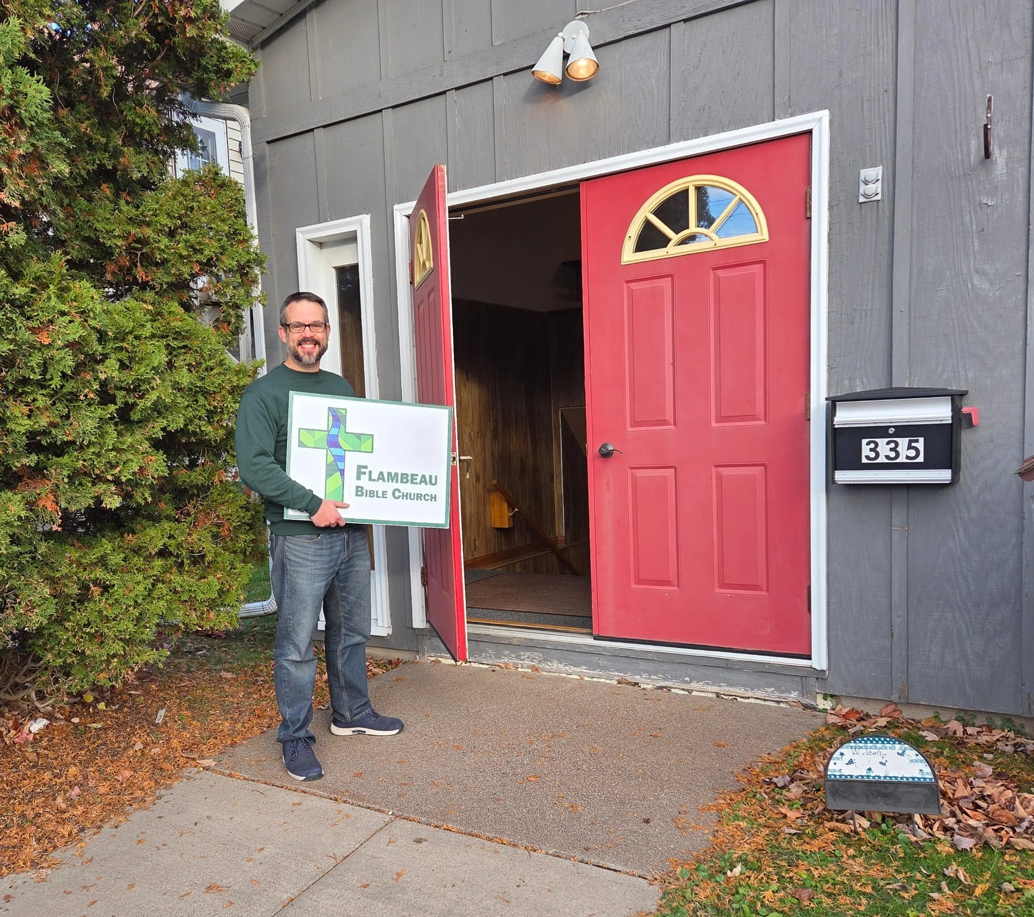 A man holding a sign stands outside the entrance of Flambeau Bible Church, with a red door, a white sign, and a house number 335, beside a bush and framed by autumn leaves.