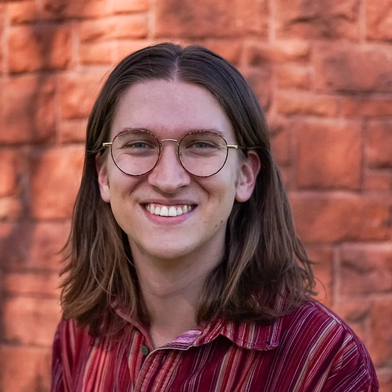 Smiling musician with long brown hair, wearing round glasses and a red striped shirt, standing in front of a brick wall.