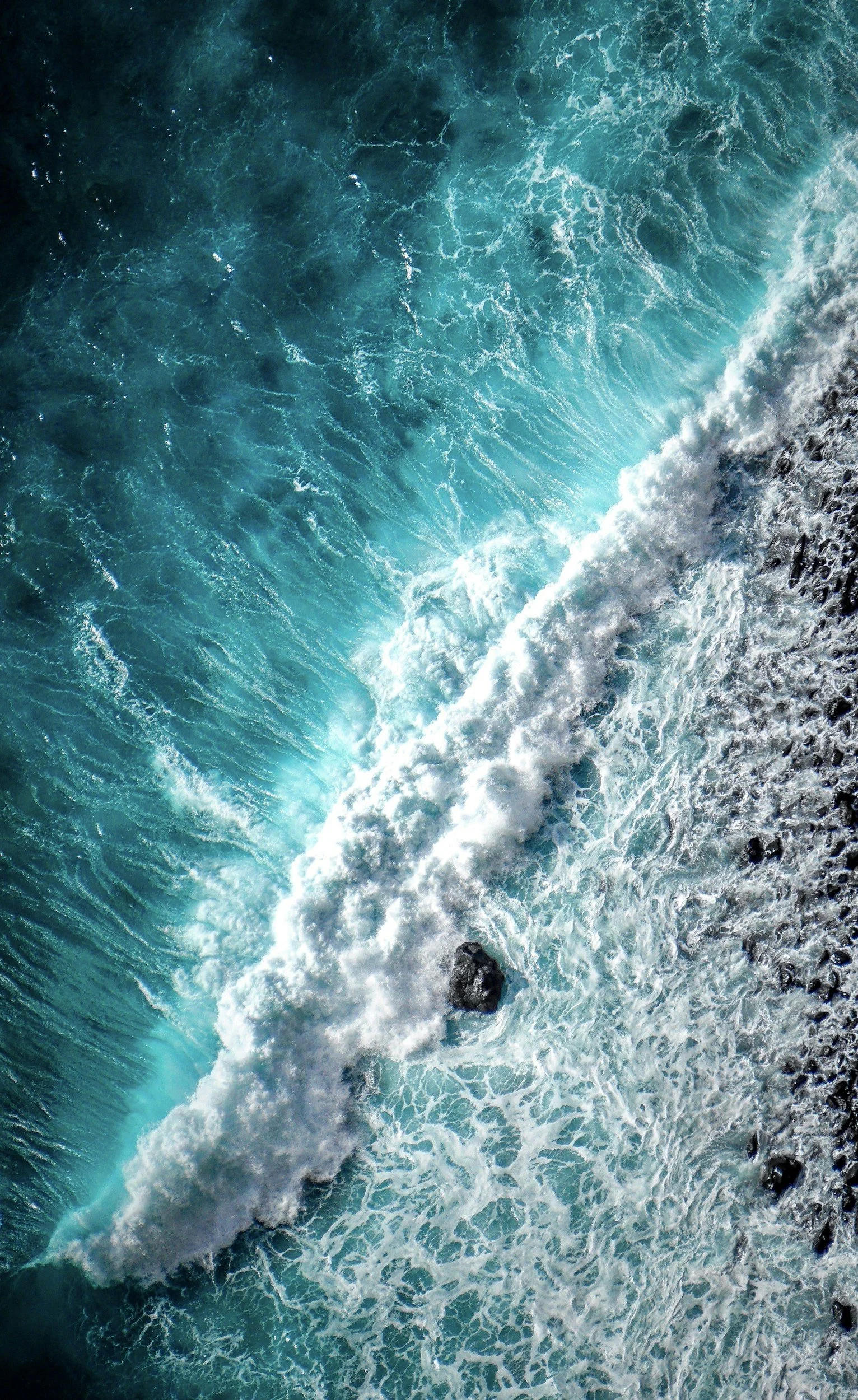 Aerial view of ocean waves crashing against rocks, turquoise water with white foam.