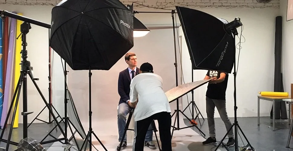 A professional photography studio setup with a Pinpoint PR client in a suit, surrounded by photography equipment including large umbrellas, and tripods, with crew adjusting a reflector.