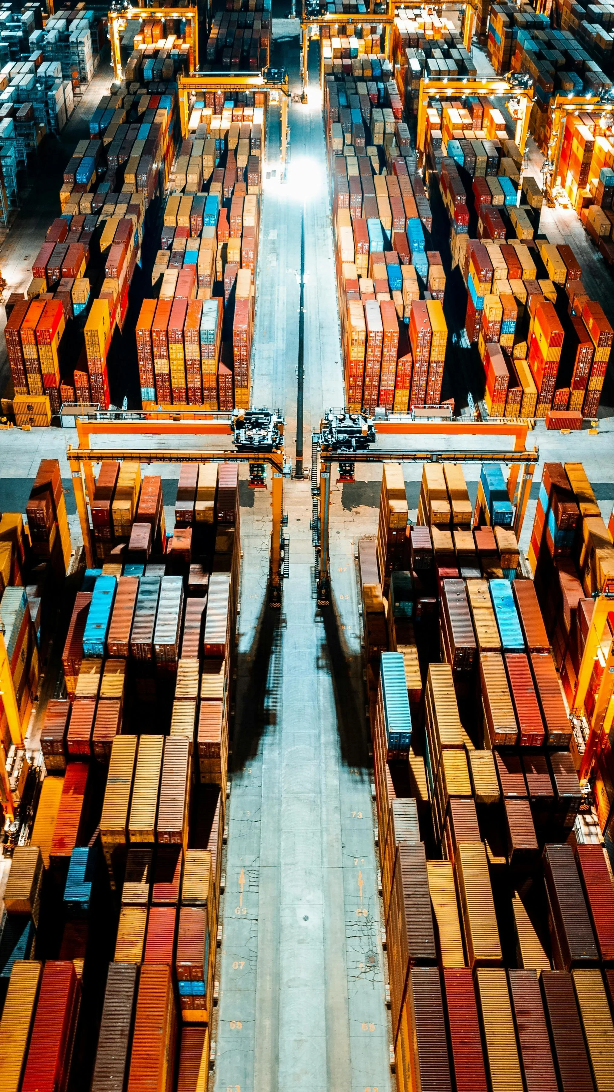 Aerial view of a container yard at night with numerous stacked shipping containers and large yellow cranes.