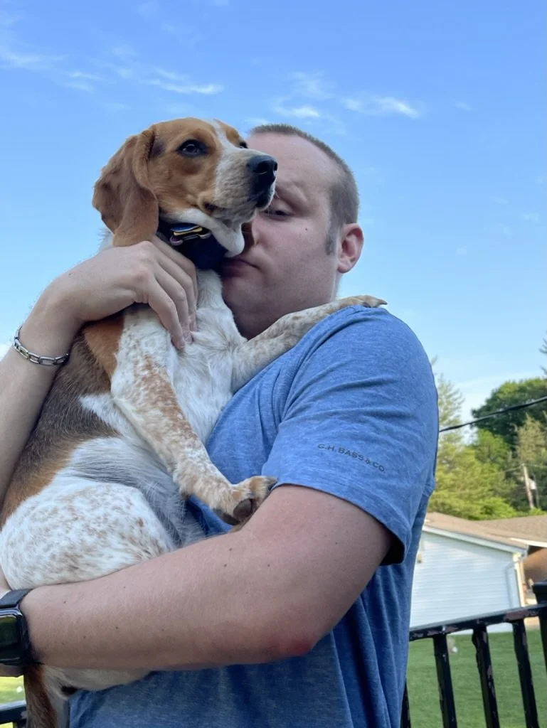 A man thriving with cancer holding a dog in his arms outside with a blue sky in the background.