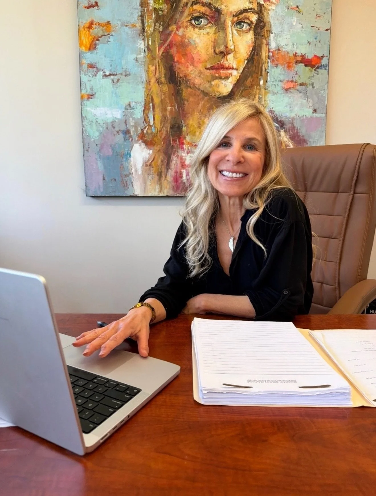 A smiling woman with long blonde hair working at her desk. She is sitting in a brown leather office chair with a stack of papers, a notebook, and a laptop on the wooden desk. Behind her is a colorful abstract portrait painting of a woman with light eyes and brown hair.