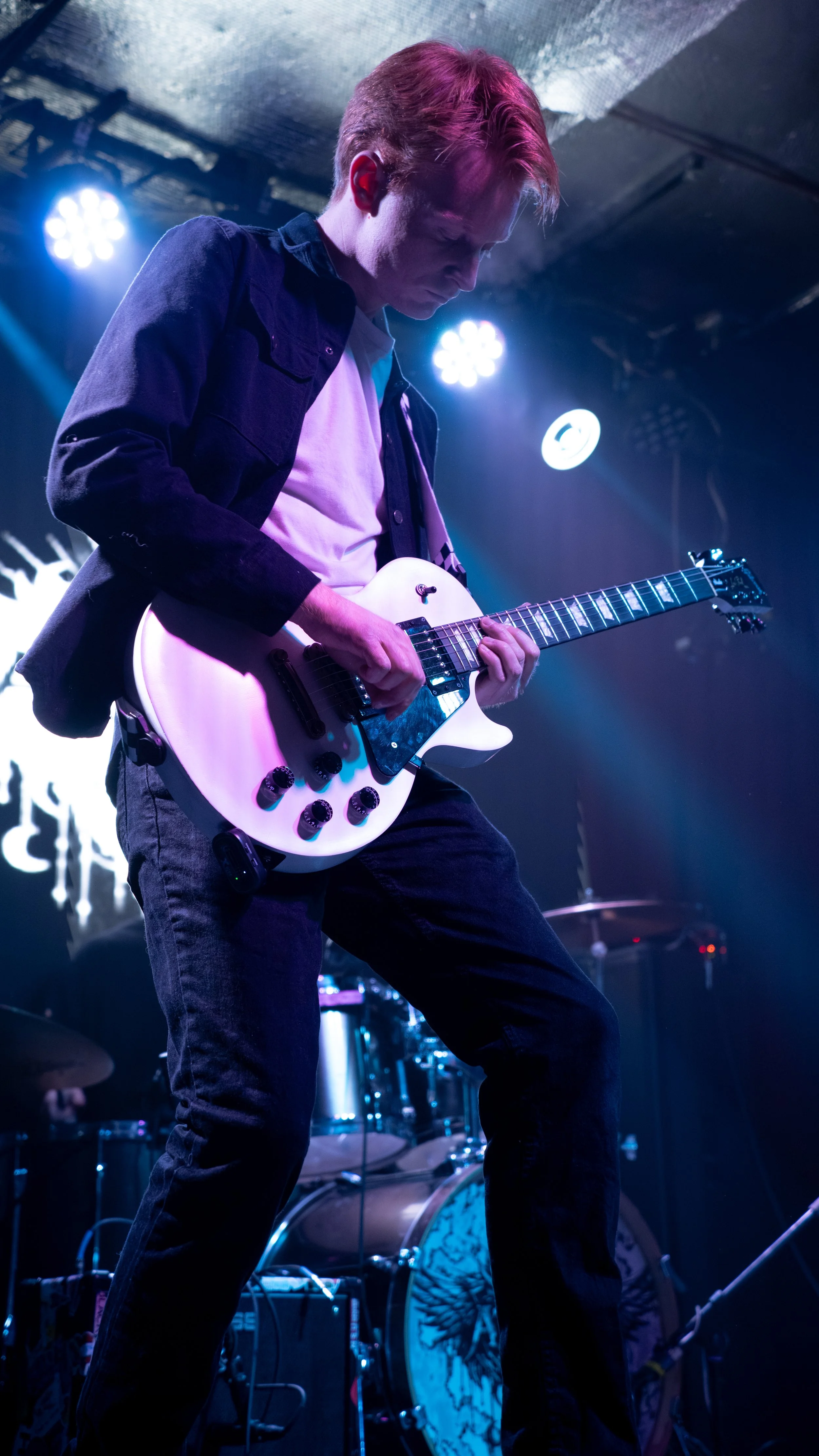 A young man playing an electric guitar on stage with stage lights and drums in the background.