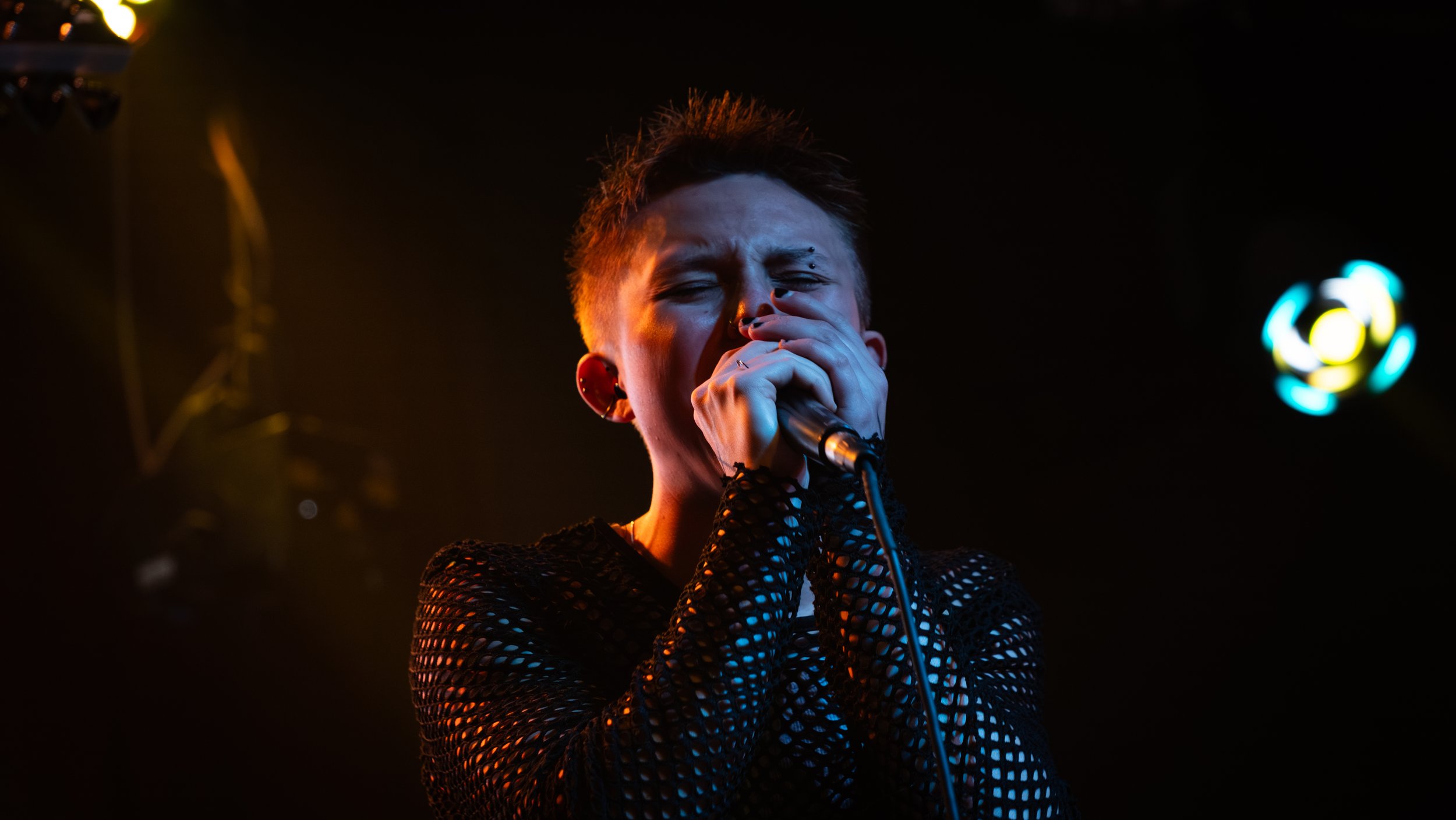 A person singing into a microphone during a performance, wearing a fishnet black top and red earrings, with a dark background and colorful stage lights.