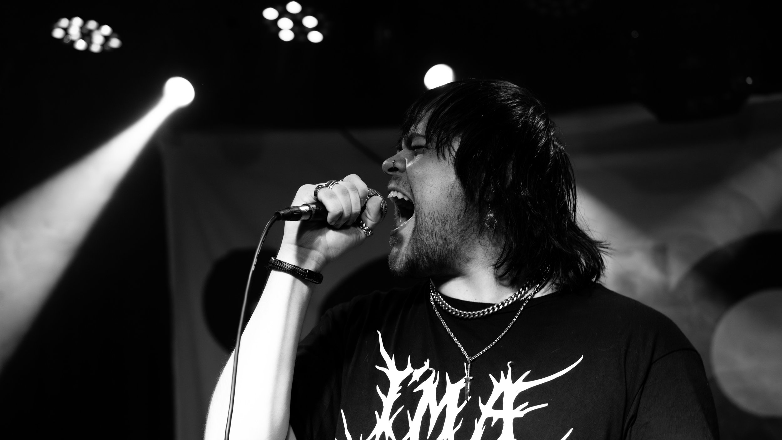 A young man singing into a microphone on stage, with long dark hair and wearing a black shirt with a white graphic. The scene is dark with stage lights overhead.