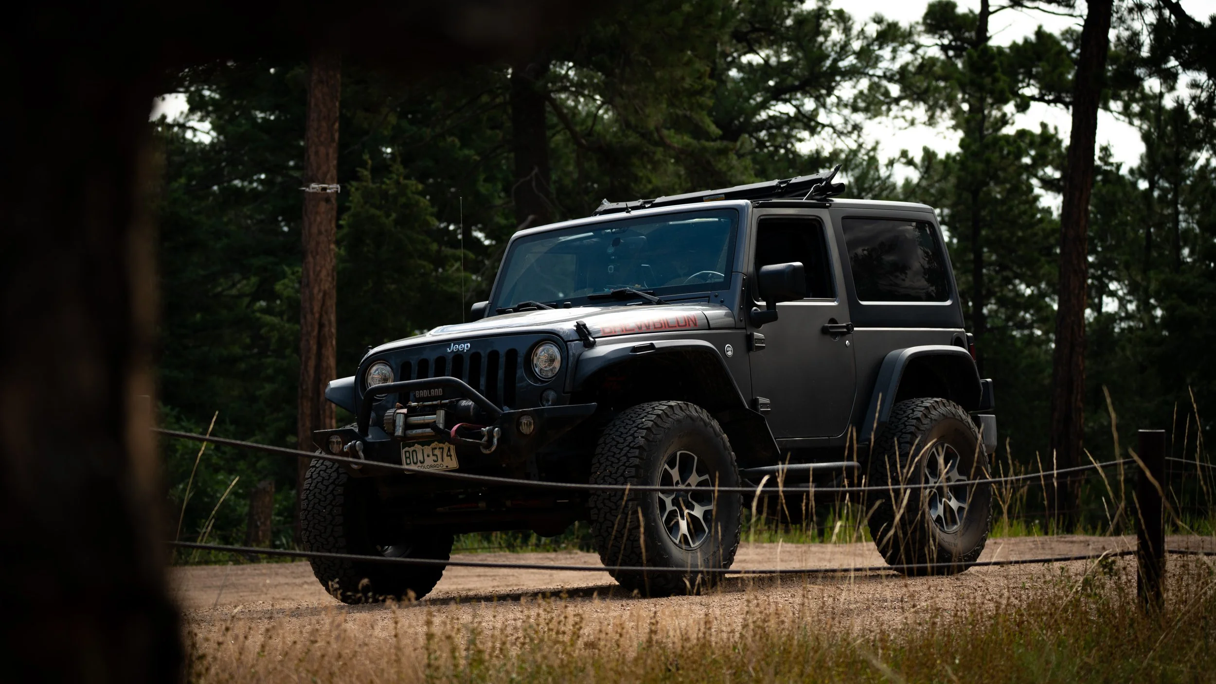 A black Jeep SUV parked on a dirt road in a wooded area with tall trees and grass in the foreground.