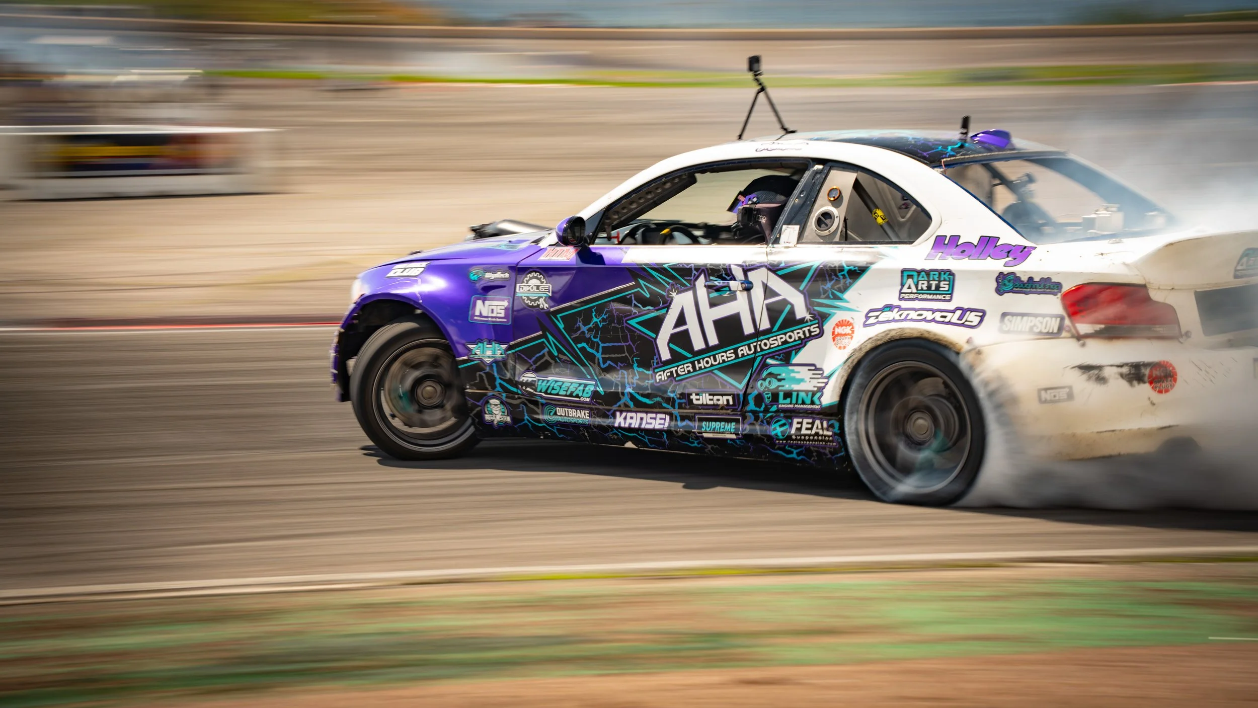 A race car speeding on a track with a blurred background, featuring a purple and white paint job with various sponsor logos and decals.